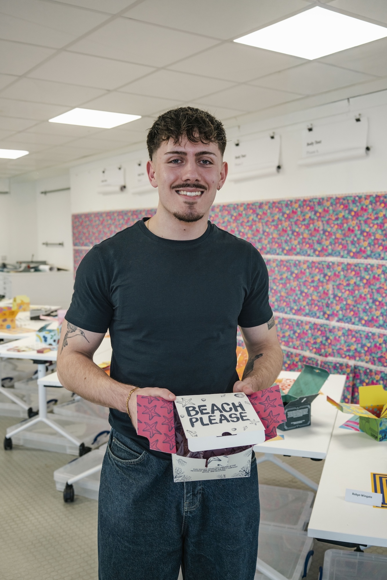 J Page holding pink and white box reading "BEACH PLEASE" in classroom with fluorescent lighting and desks.