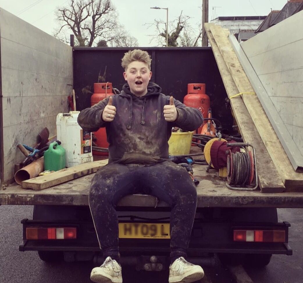 A young man in a dark hoodie and work trousers sits on the back of a flatbed truck, giving two thumbs up, with gas cylinders, tools and wooden planks loaded behind him.