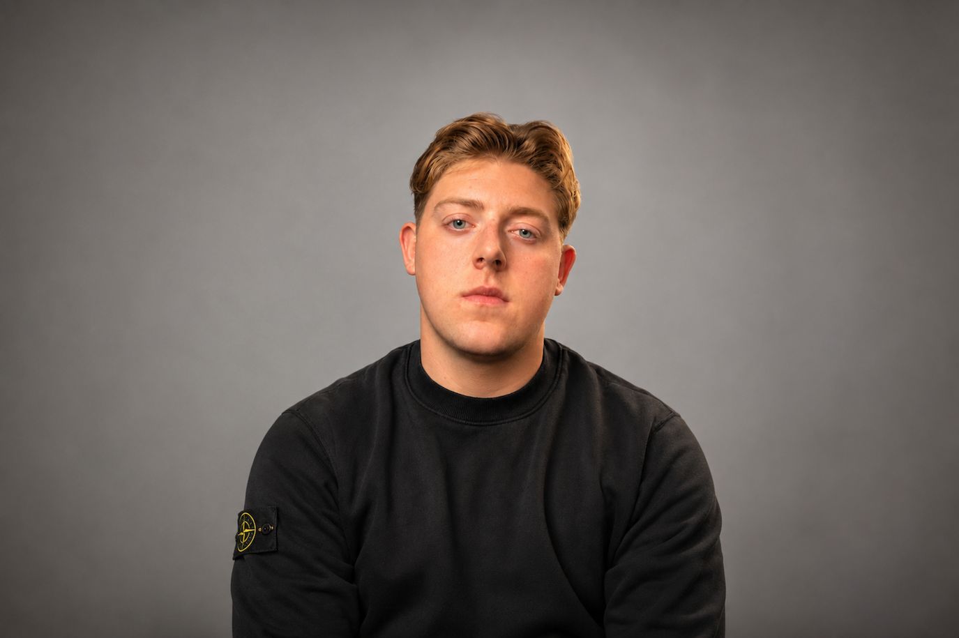A young man in a black sweatshirt against a plain grey studio background.