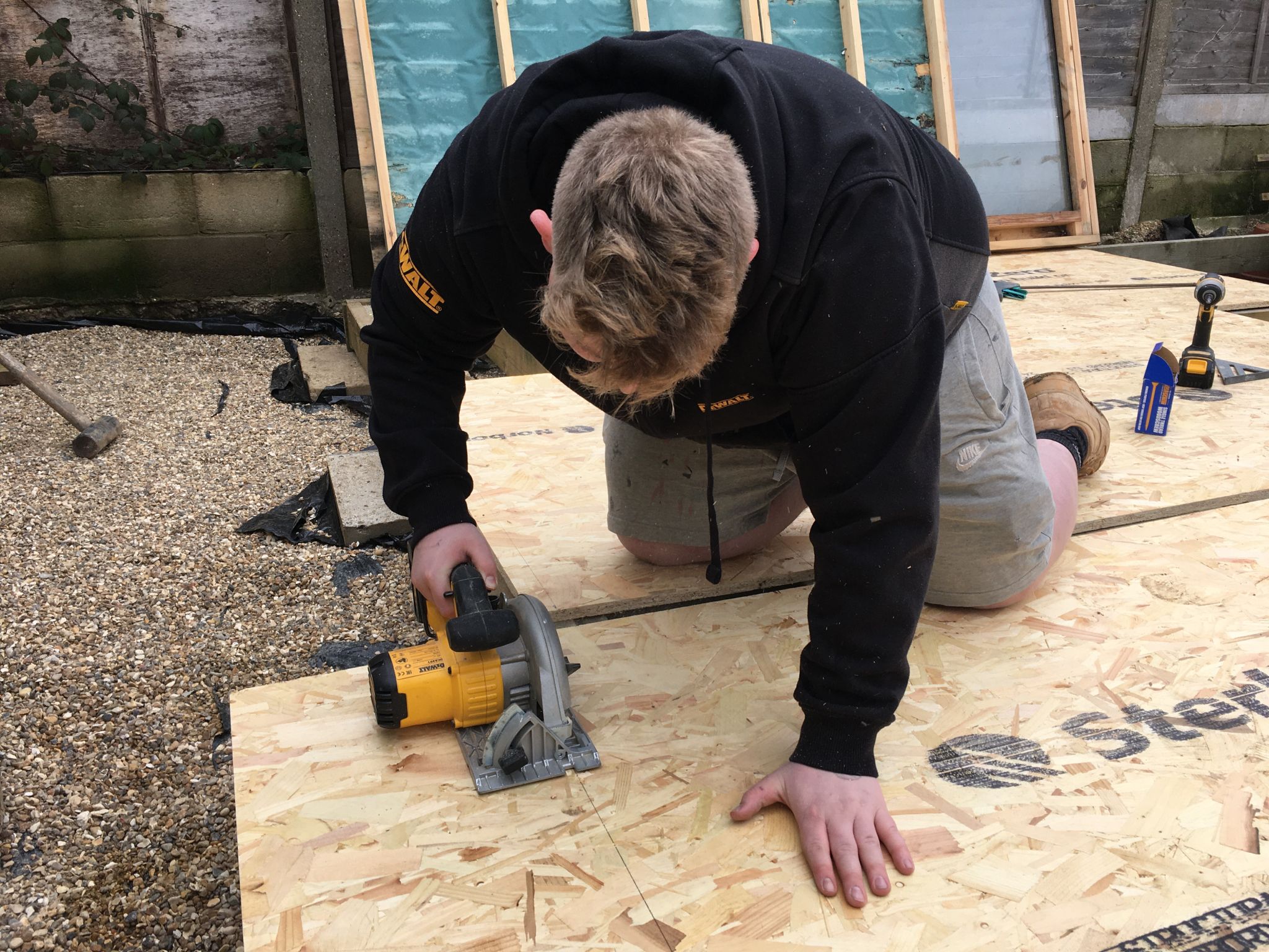 A man in a black hoodie kneels on plywood, using a circular saw to cut a sheet on an outdoor work site, with gravel, timber and tools in the background.