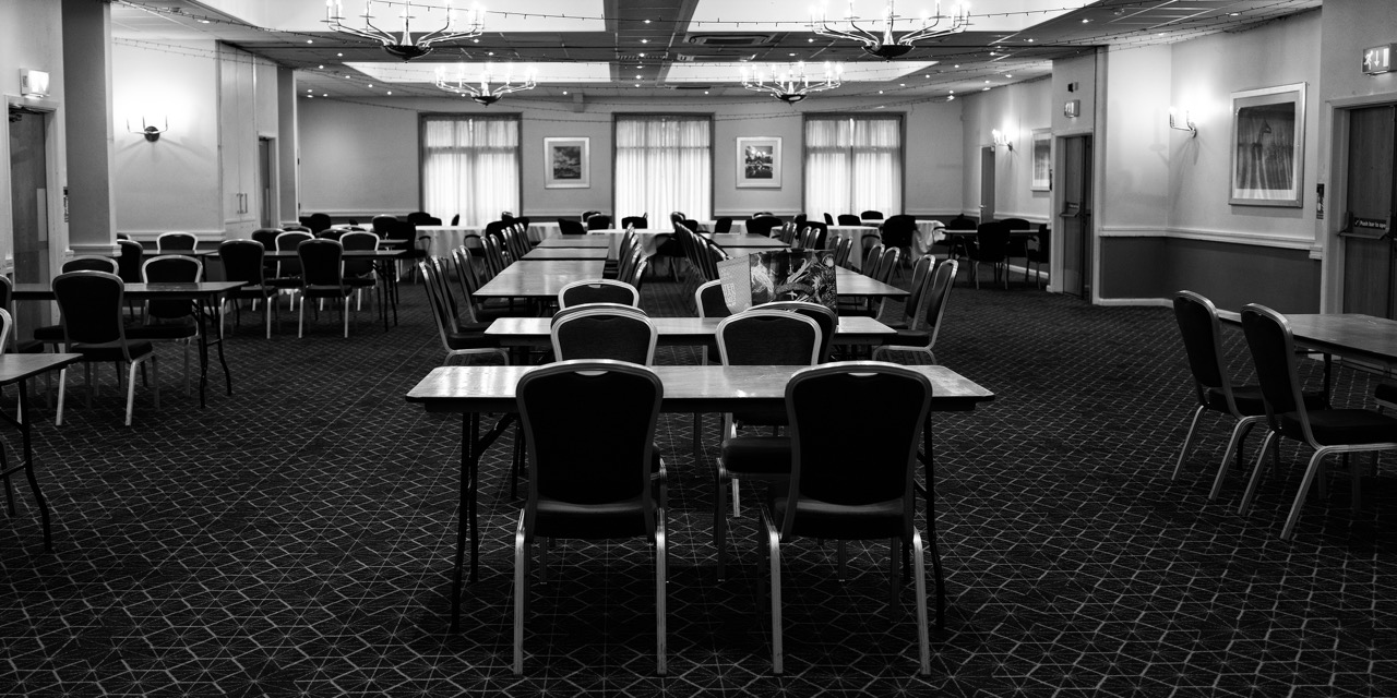 An empty conference room with rows of chairs and tables arranged for a meeting, with windows and framed pictures on the back wall.