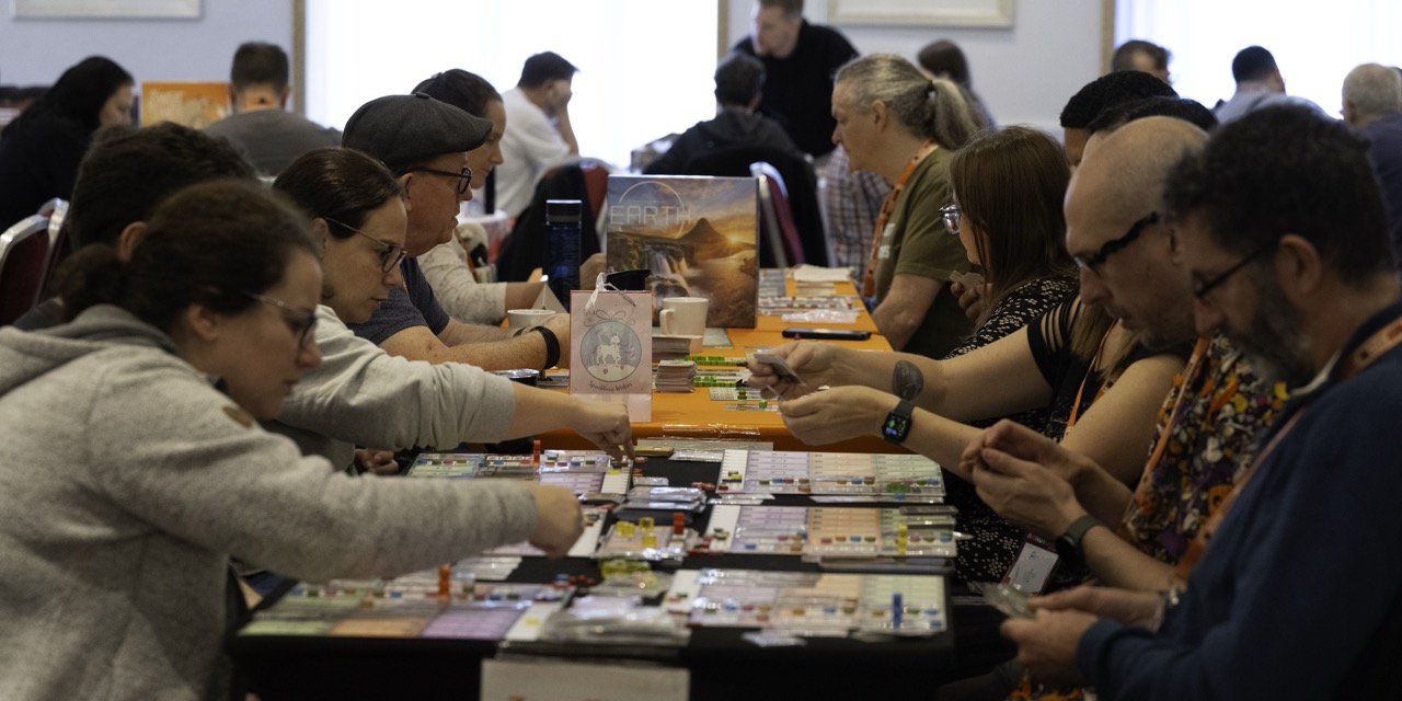 A group of people seated at a long table browse books and small items at an indoor market or fair, with displays arranged down the centre.