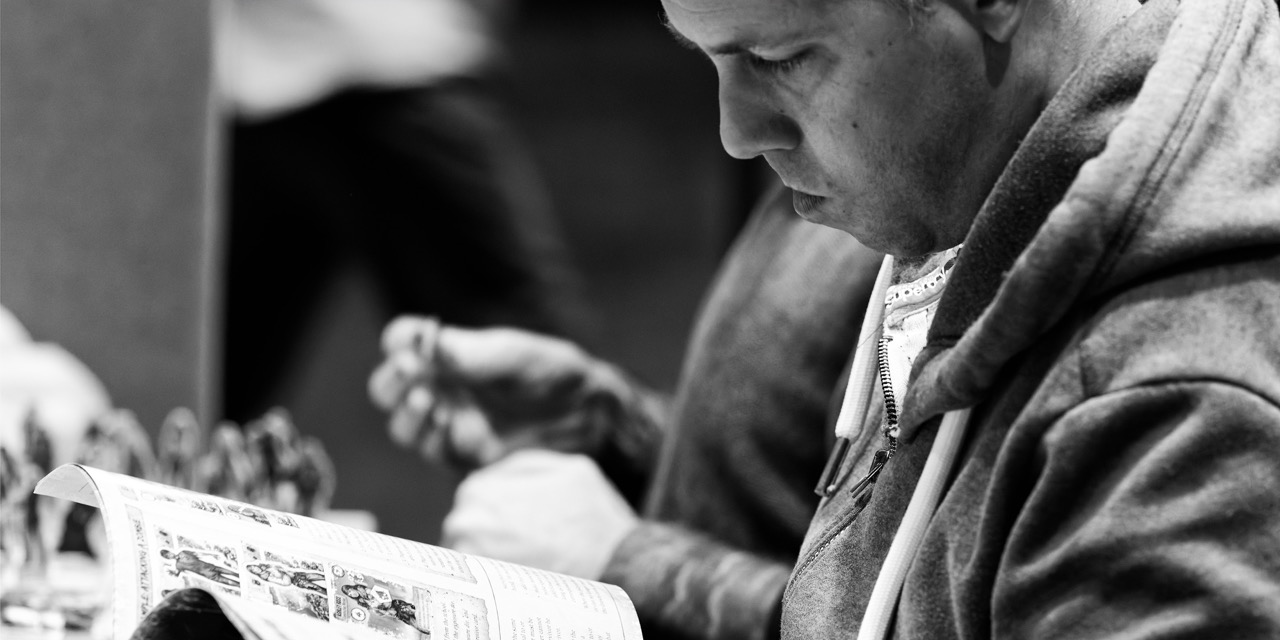 A man in a hooded sweatshirt reads a magazine at a table, with another person blurred in the background.