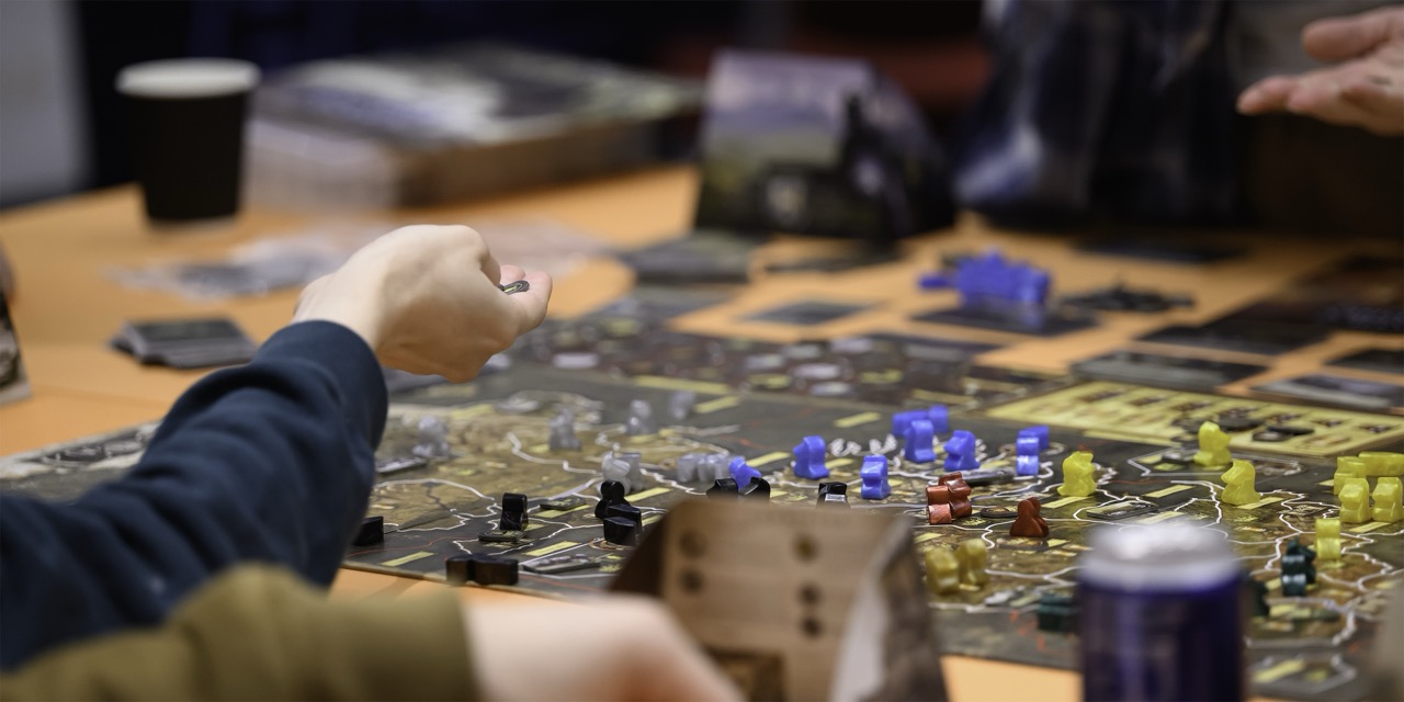 Players move coloured pieces on a tabletop board game, with a coffee cup and drinks on the wooden table.