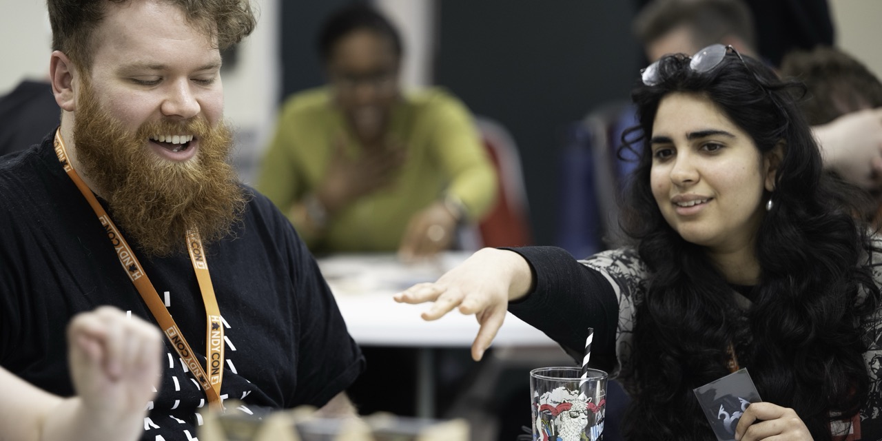 A bearded man wearing a lanyard sits beside a woman with long dark hair and sunglasses on her head, reaching across a table with a drink in the foreground and blurred people behind them.
