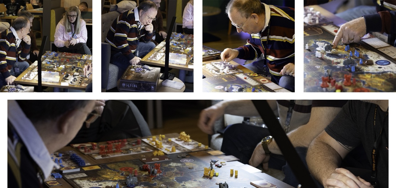 A group of people play a tabletop board game around a large table, with small game pieces and illustrated boards spread across the surface.