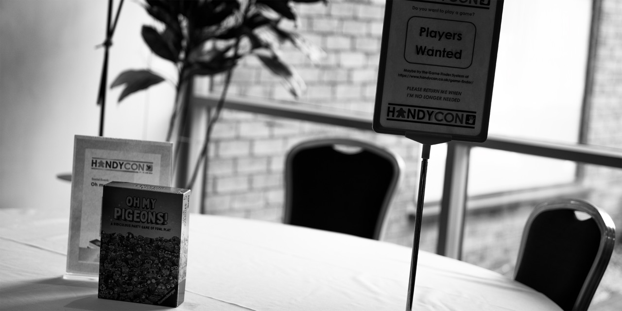 A table display with a standing sign reading “Players Wanted”, a book cover titled “Dirty Pigeons”, and a menu holder in a café by a brick wall and window.
