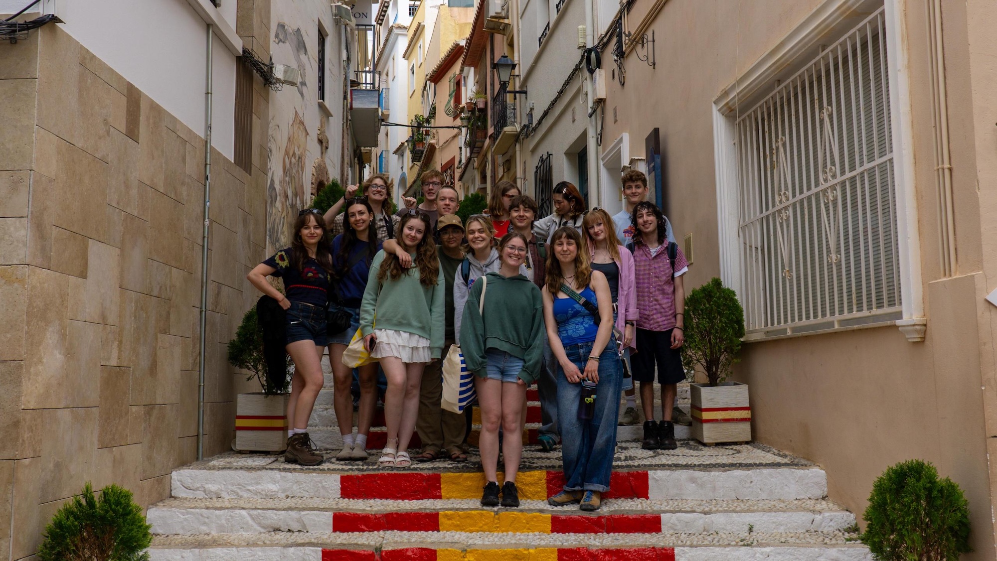 A group of young people stands on outdoor steps painted with red and yellow stripes, between narrow buildings with windows and potted shrubs.