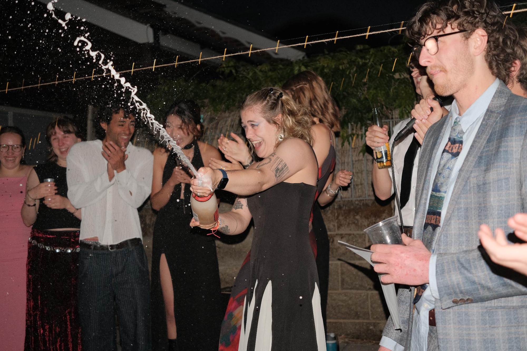 A woman in a black dress opens a champagne bottle at an outdoor party, with guests standing nearby and a spray of foam in the air.
