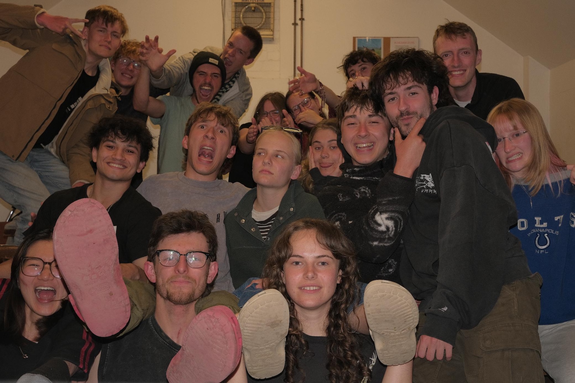 A group of young adults pose indoors on a staircase, with several people leaning close together and a pair of pink and white shoe soles held towards the camera.