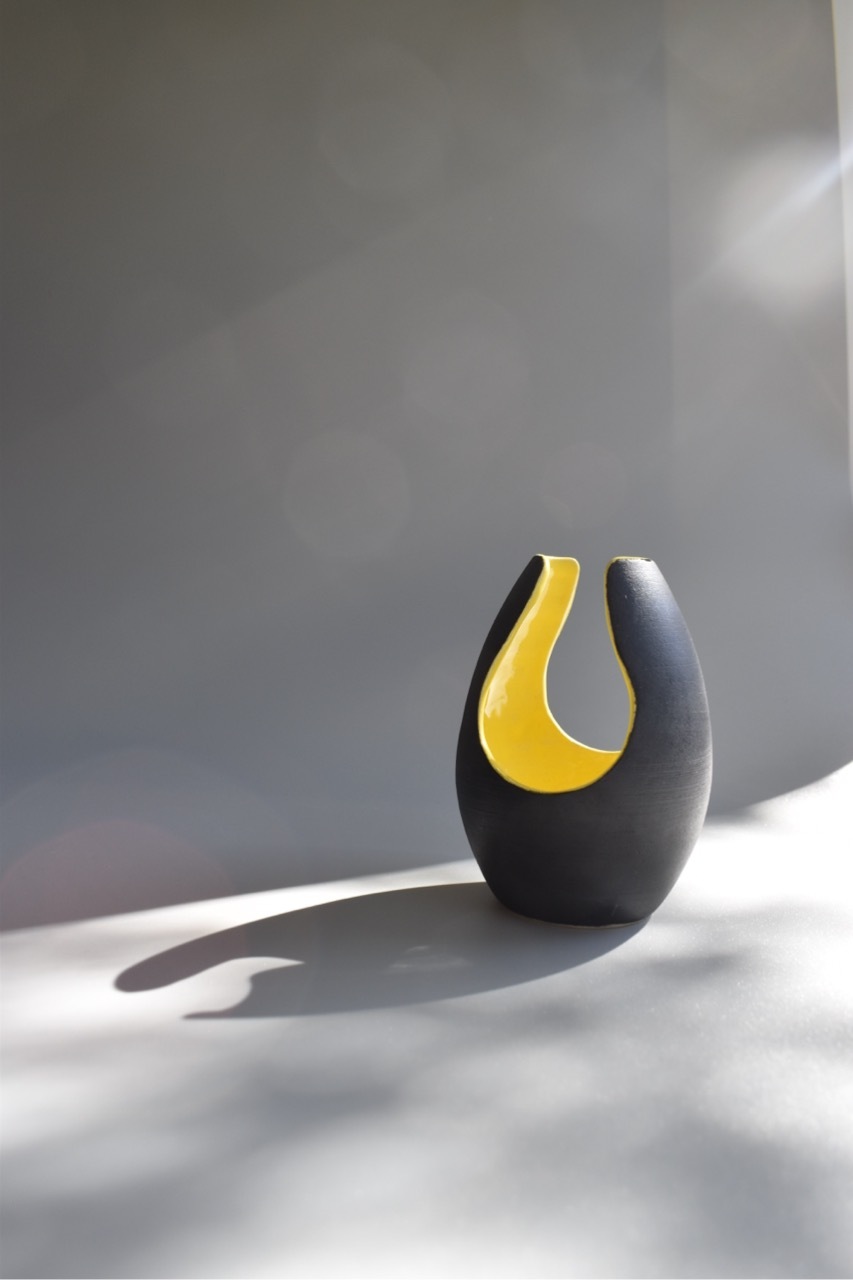 Black ceramic vase with yellow interior curve, casting shadow on white surface against blurred grey background.