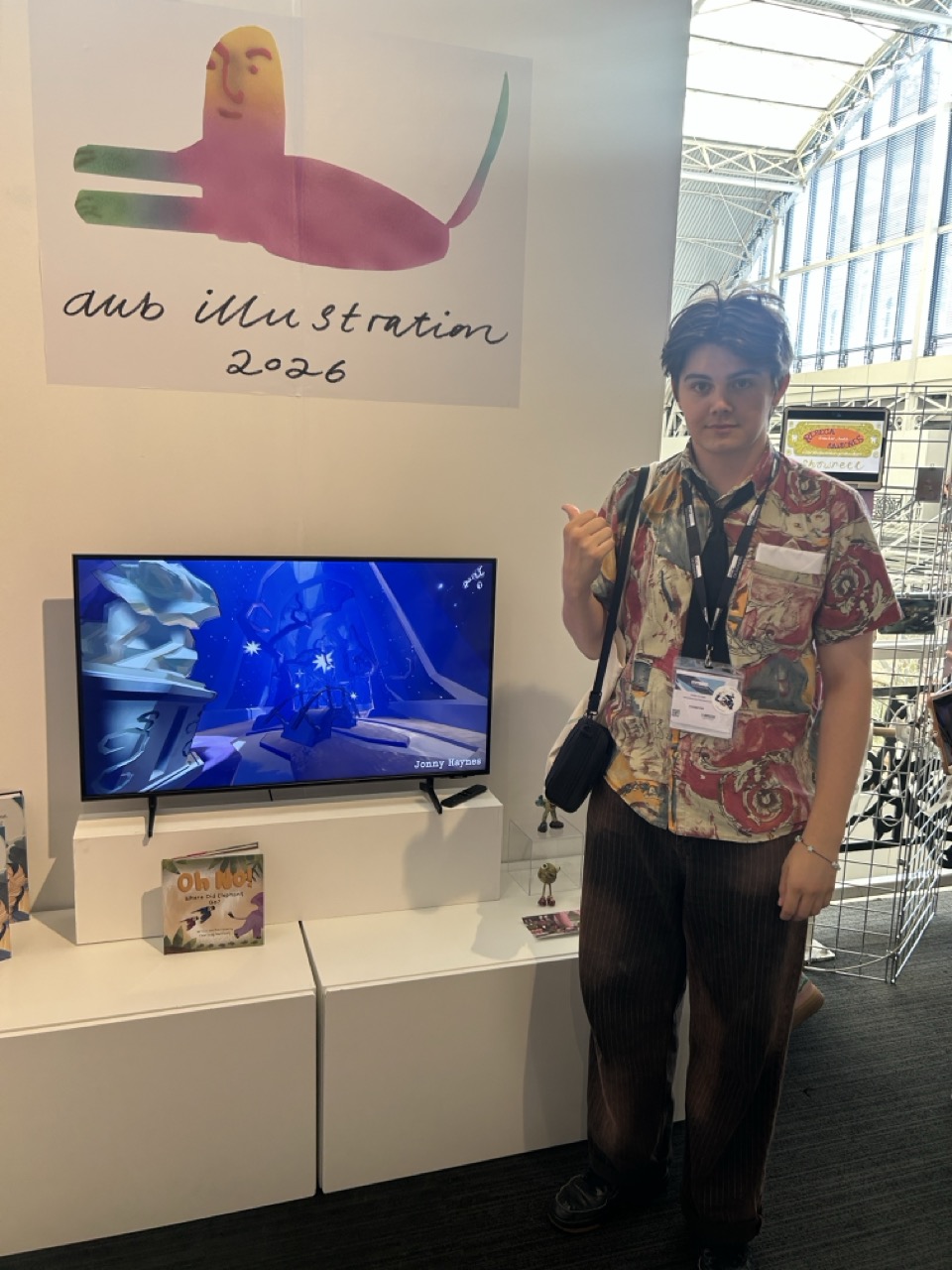A person wearing a patterned shirt and lanyard stands beside a wall-mounted screen at a trade fair booth, with an 'aus illustration 2026' sign above.