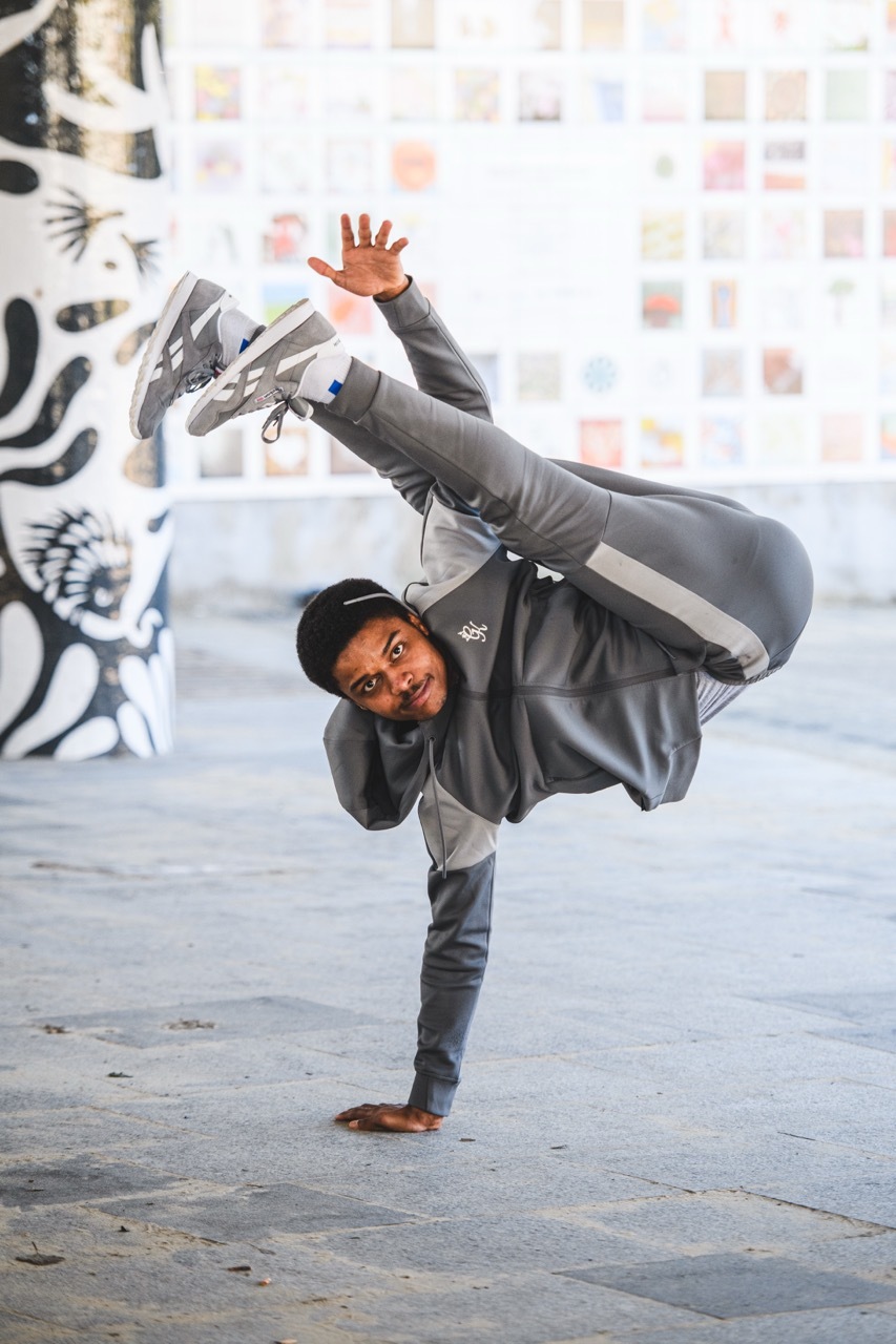 Dancer performing one-handed freeze move, balancing on left hand with legs raised, wearing grey tracksuit and black cap.