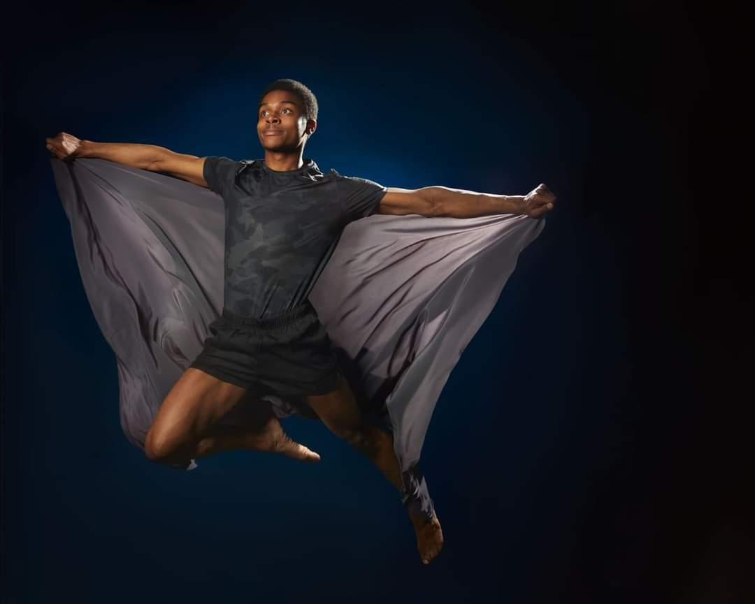 Male dancer mid-leap with arms outstretched holding flowing grey fabric against dark blue background with dramatic lighting.