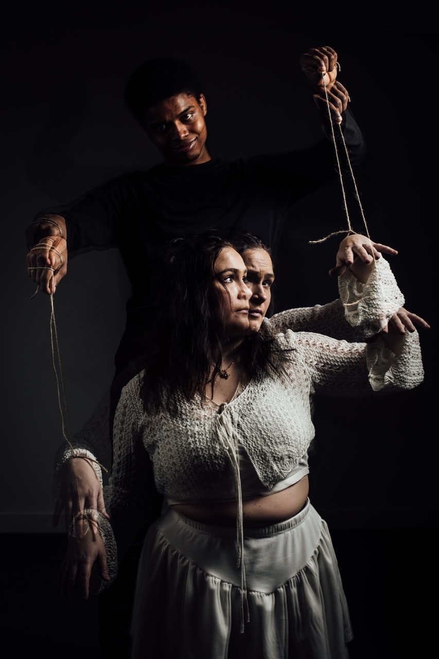 Woman in cream cardigan and white skirt manipulated by a man and multiple hands holding strings like a marionette against black background.