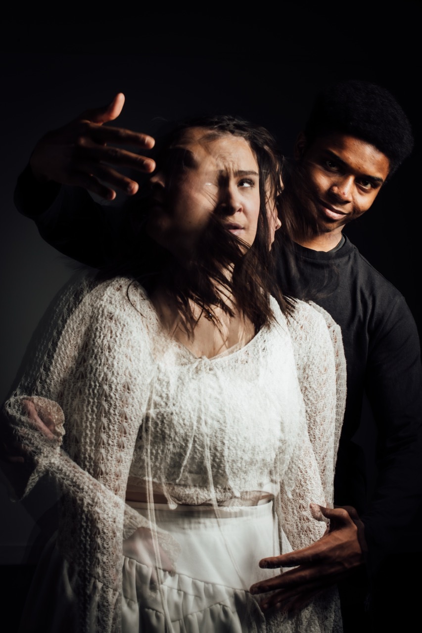 Woman in white dress shielding face with hand whilst man in dark clothing stands behind her against black background.