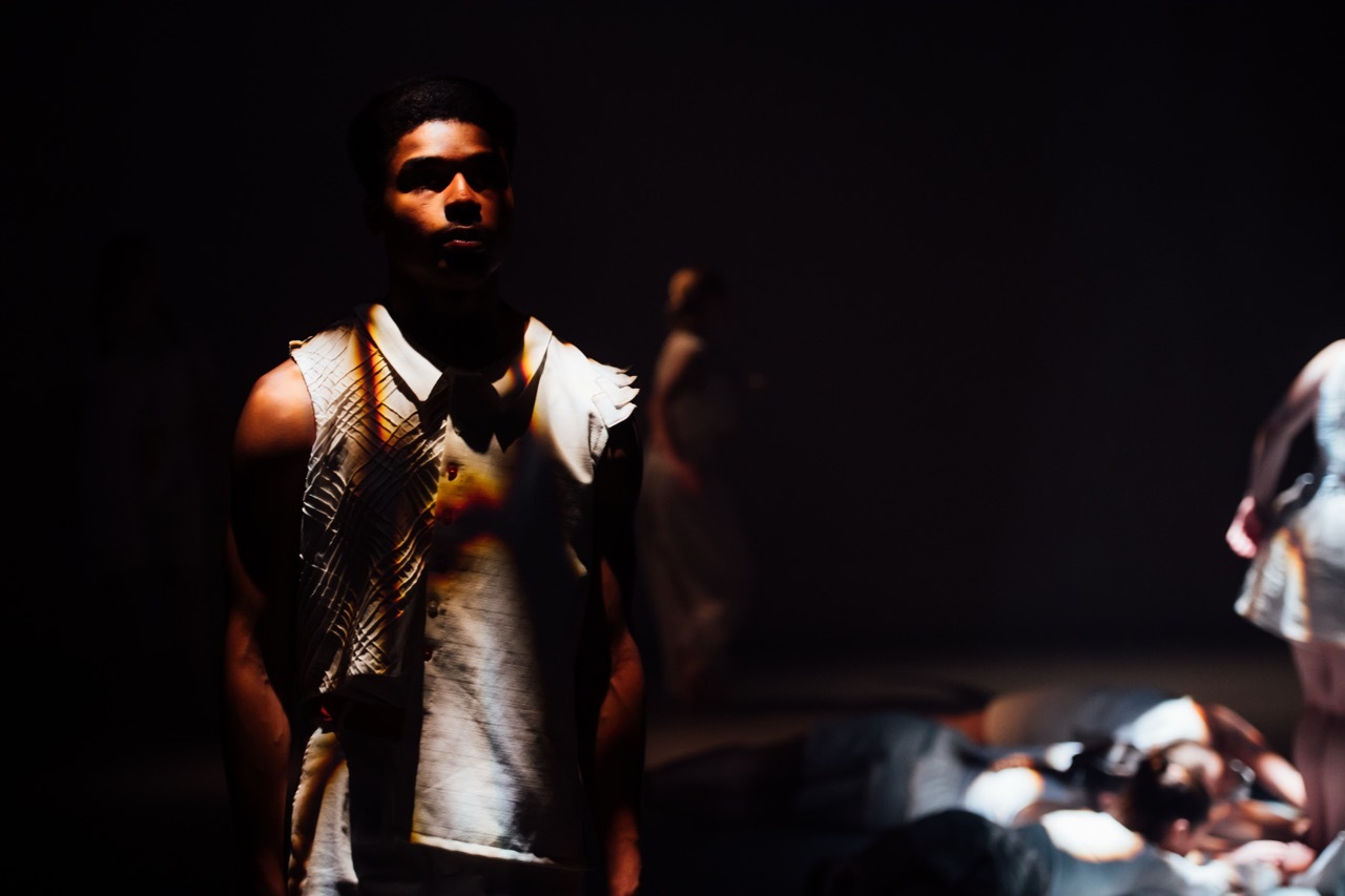 Man in patterned waistcoat and white shirt performing on dark stage with dramatic lighting and blurred figures in background.