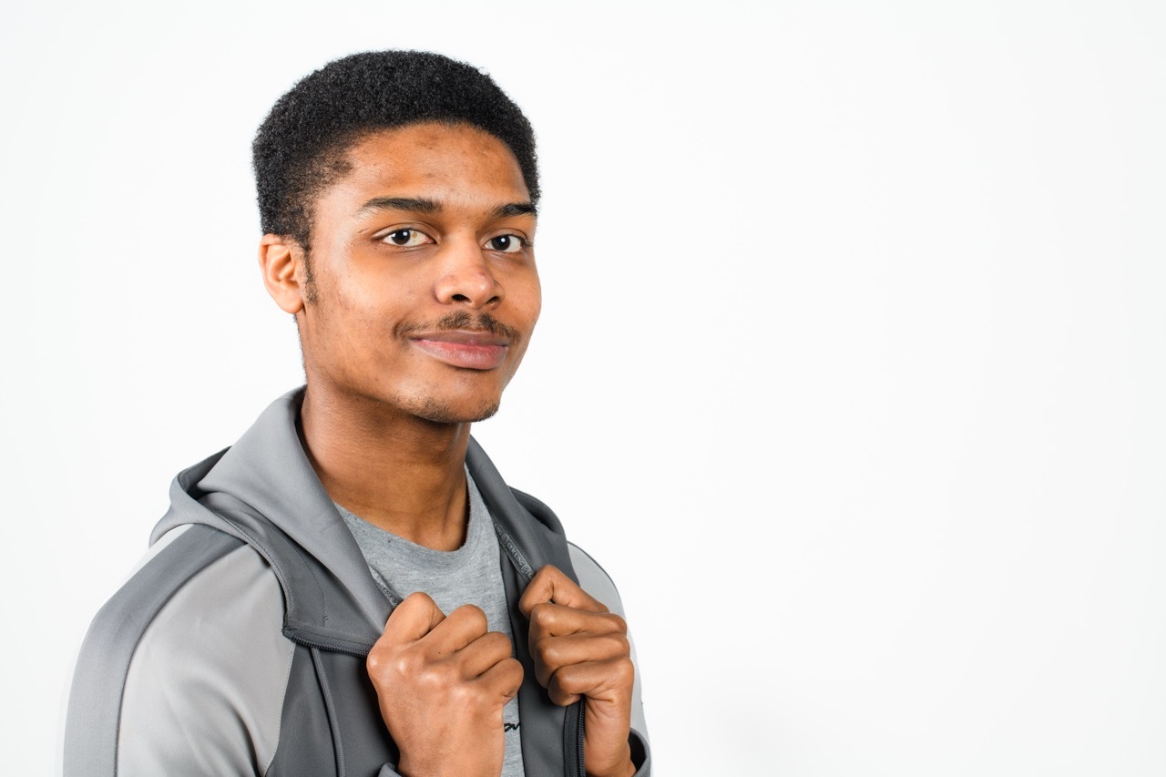 Young man with short hair wearing grey hoodie, holding jacket collar, smiling at camera against white background.