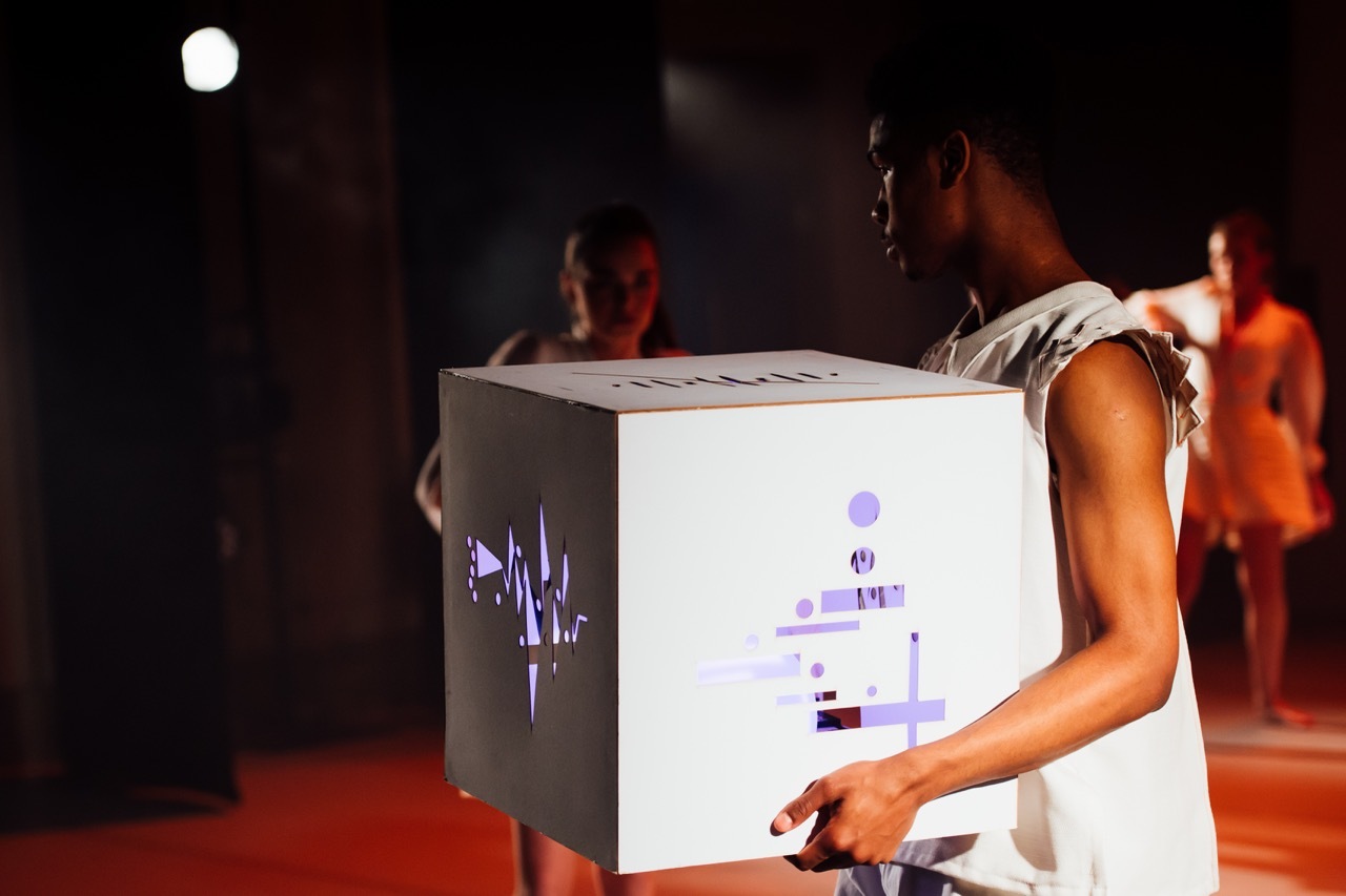 Person interacting with white cube with blue glowing light through damage and tears, dark stage with orange lighting and people in background.