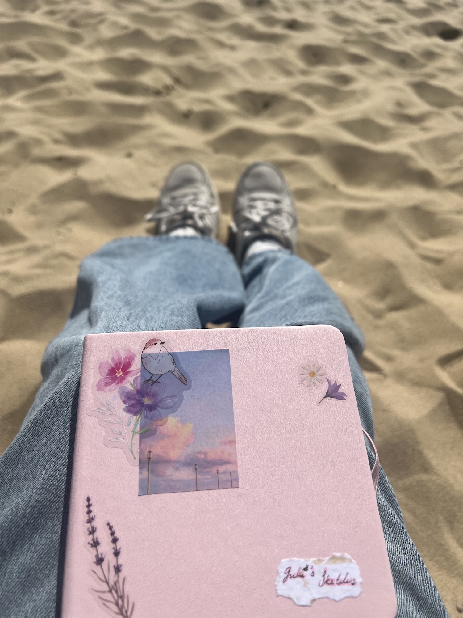 Close-up of a person's lap and a pink sketchbook, sitting on a sandy beach.