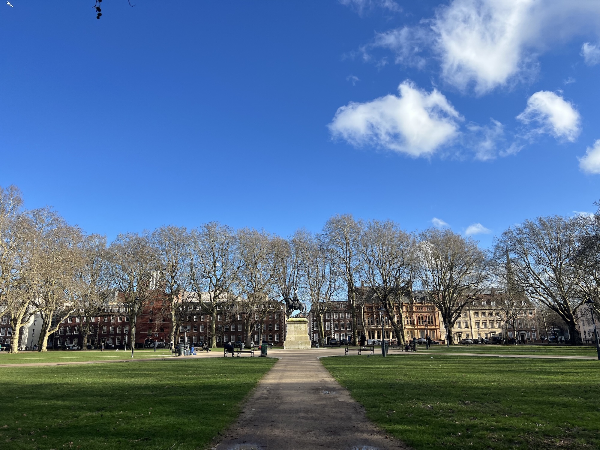 Large green lawn with central path leading to statue, surrounded by Georgian terraced buildings under bright blue sky with white clouds.