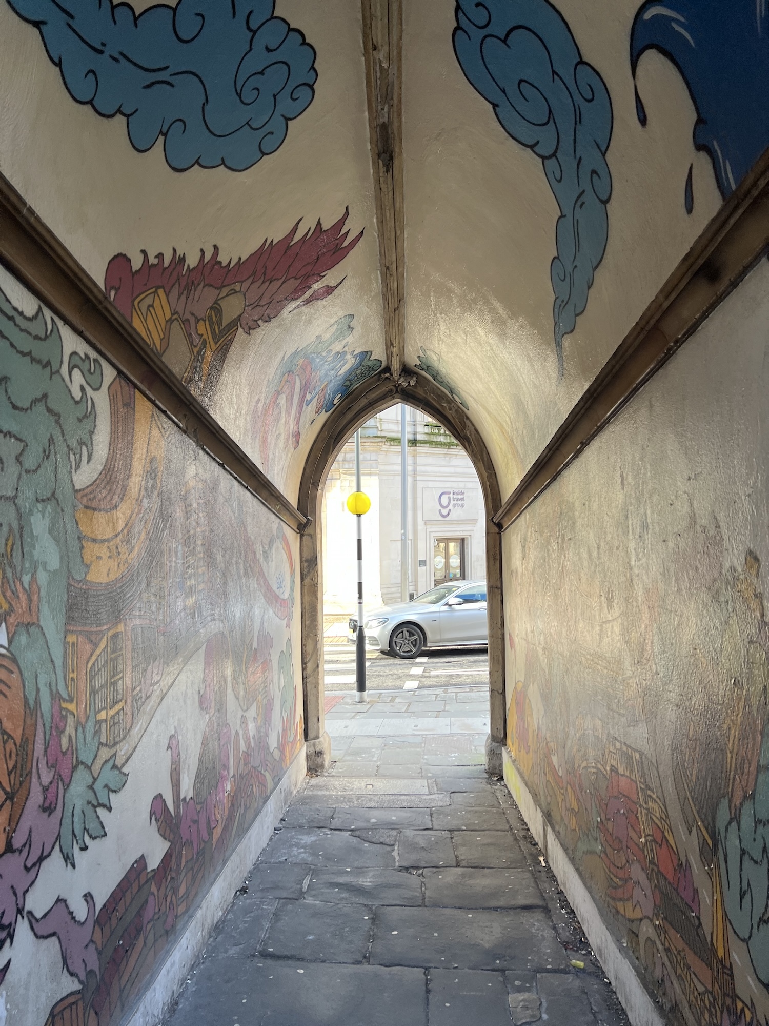 Stone archway tunnel with colourful murals on walls and ceiling featuring blue clouds, purple and green shapes, leading to street view.