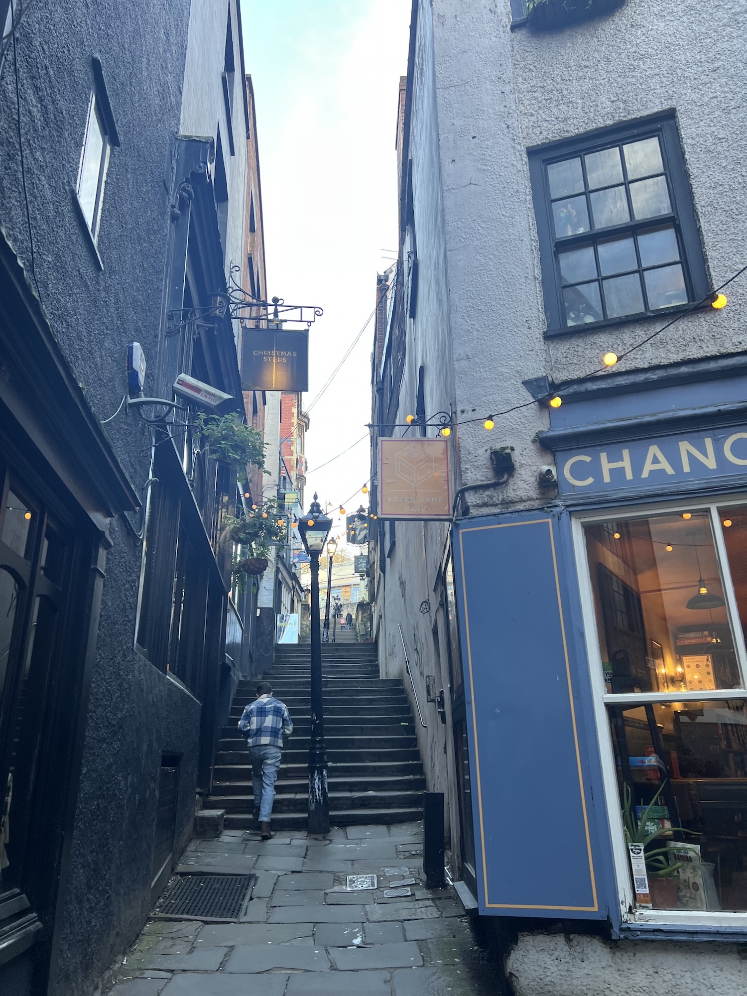 Narrow cobbled alleyway with stone steps leading upward between old buildings. Blue shopfront on right, string lights overhead, grey stone walls.