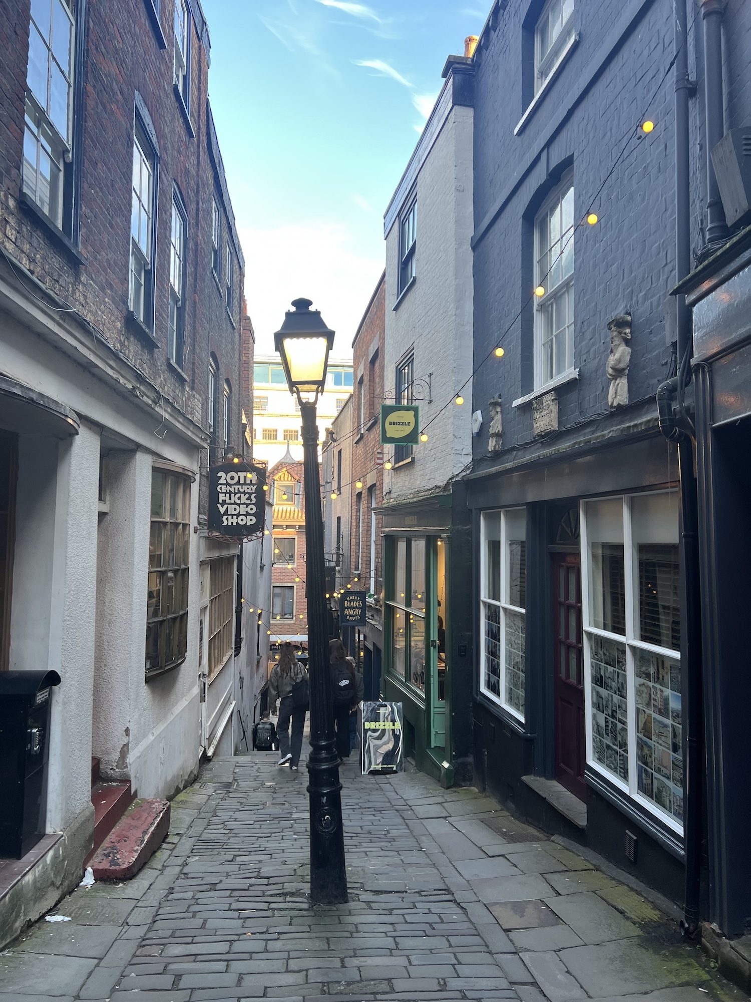 Narrow cobbled alley between multi-storey buildings with shop signs, street lamp, and stone paving under blue sky with clouds.