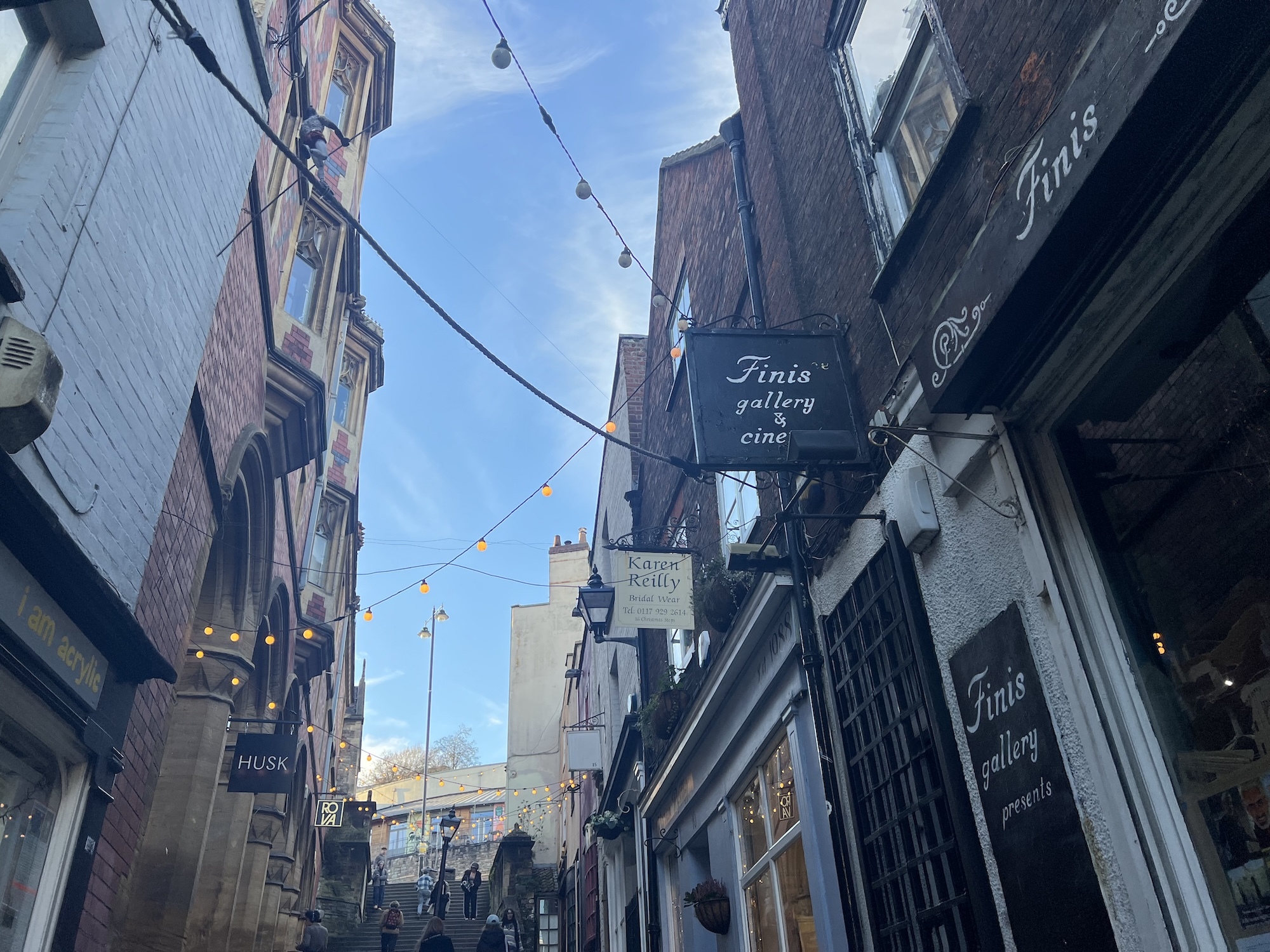 Narrow street with brick and white buildings, overhead cables, and dark shop signs including "Finto gallery" under blue sky.