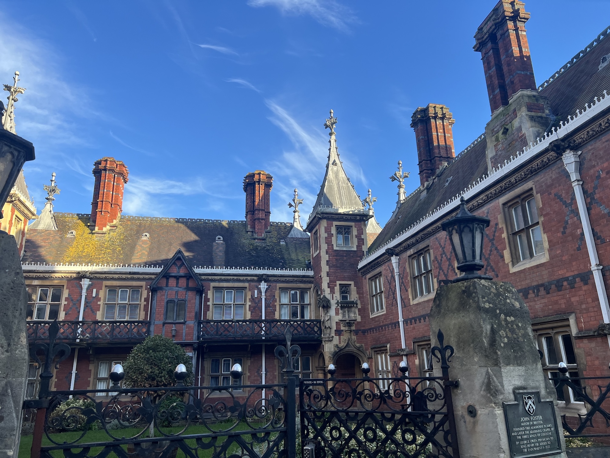 Red brick Victorian mansion with ornate chimneys, decorative stonework, and iron gates under bright blue sky with white clouds.