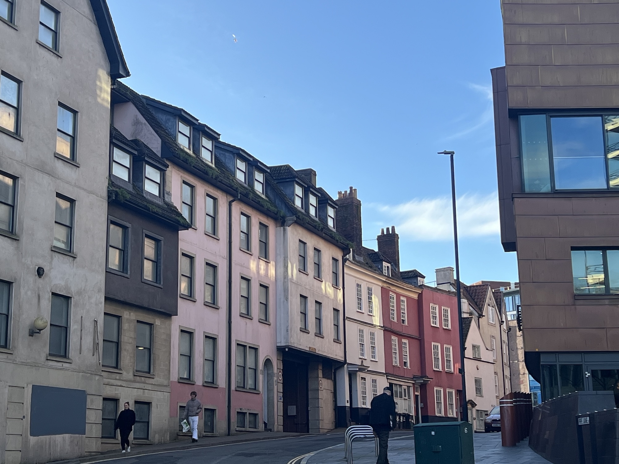 Row of colourful terraced houses in pink, yellow, and red with modern grey buildings on either side under blue sky with clouds.
