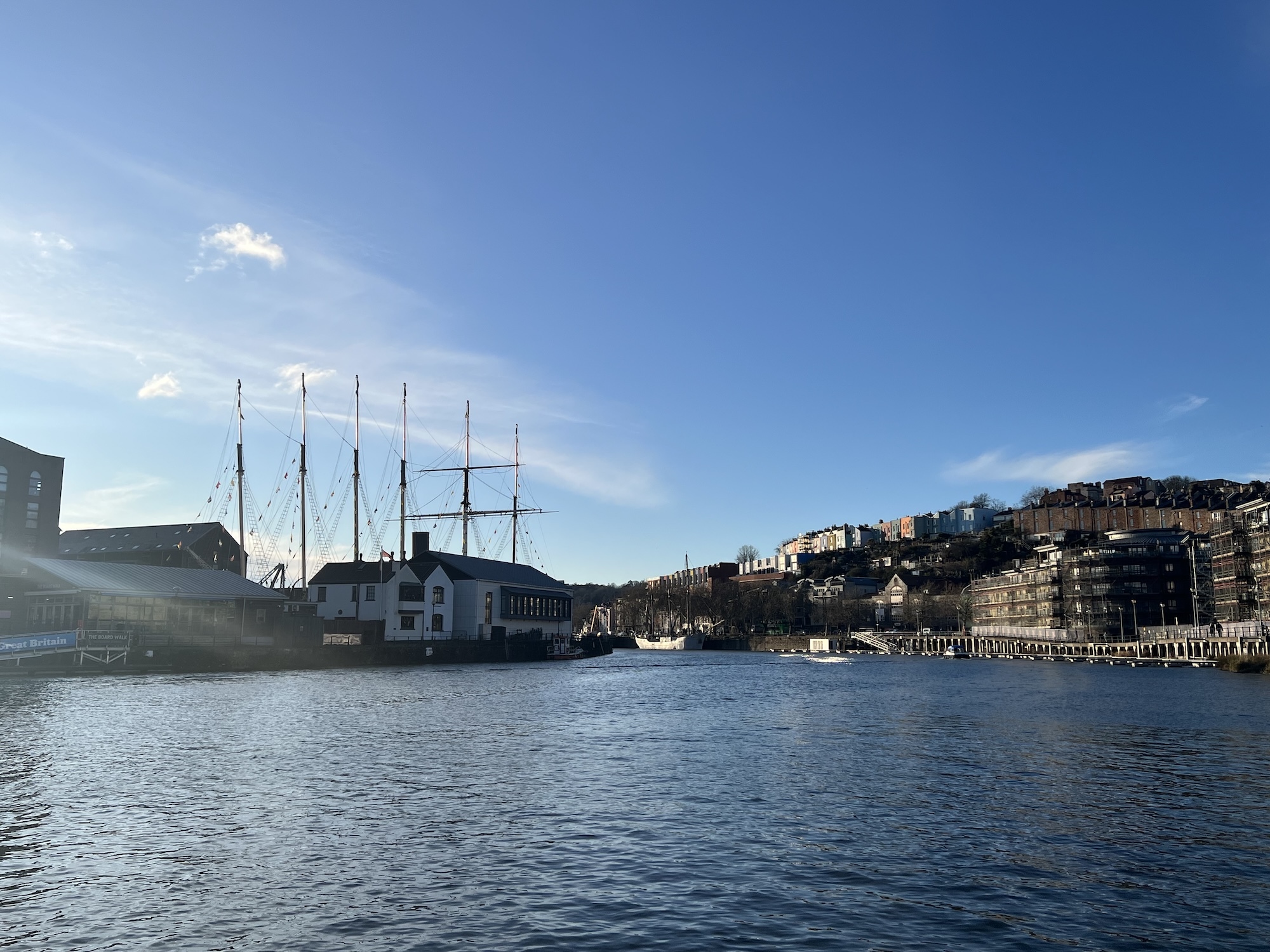 Harbour with tall sailing ship moored near white buildings, hillside town with coloured houses rising beyond, blue water and sky.
