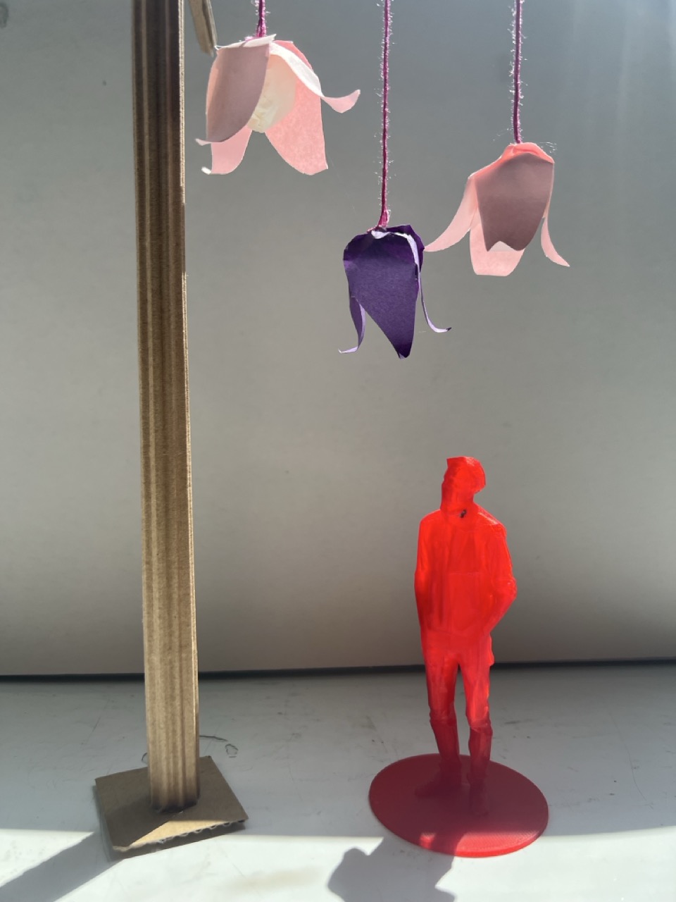 A red cut-out figure stands on a circular base, positioned below three hanging paper flower decorations in pink and purple. The figure is placed on a white surface with a vertical brown paper structure to the left.