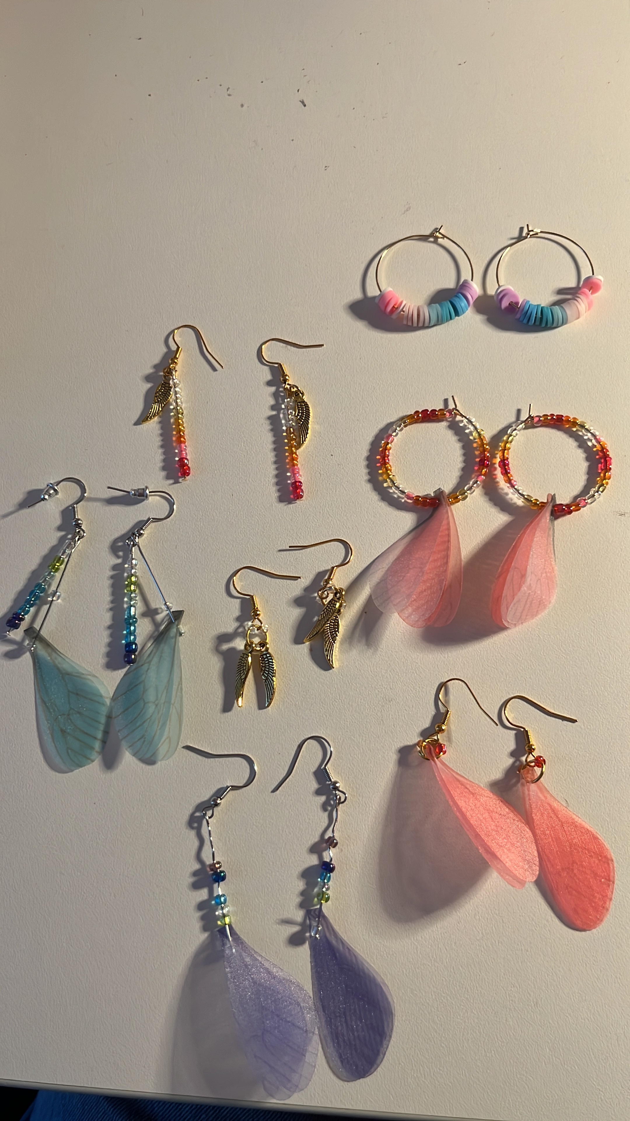 Collection of handmade earrings with colourful feathers, beaded hoops, and chain details arranged on white surface.