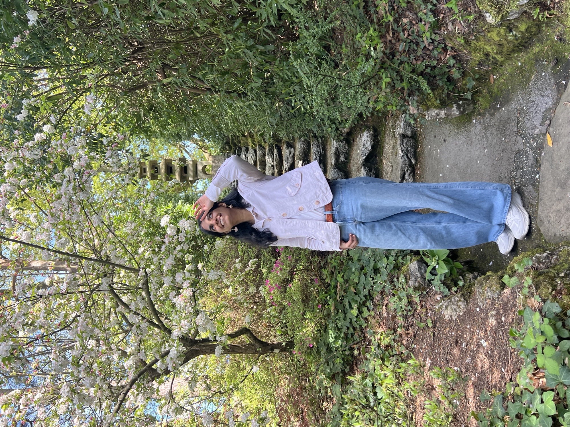 A woman in a white blouse and blue jeans stands on a stone path in a leafy garden, beside a wall covered with greenery and flowering plants.