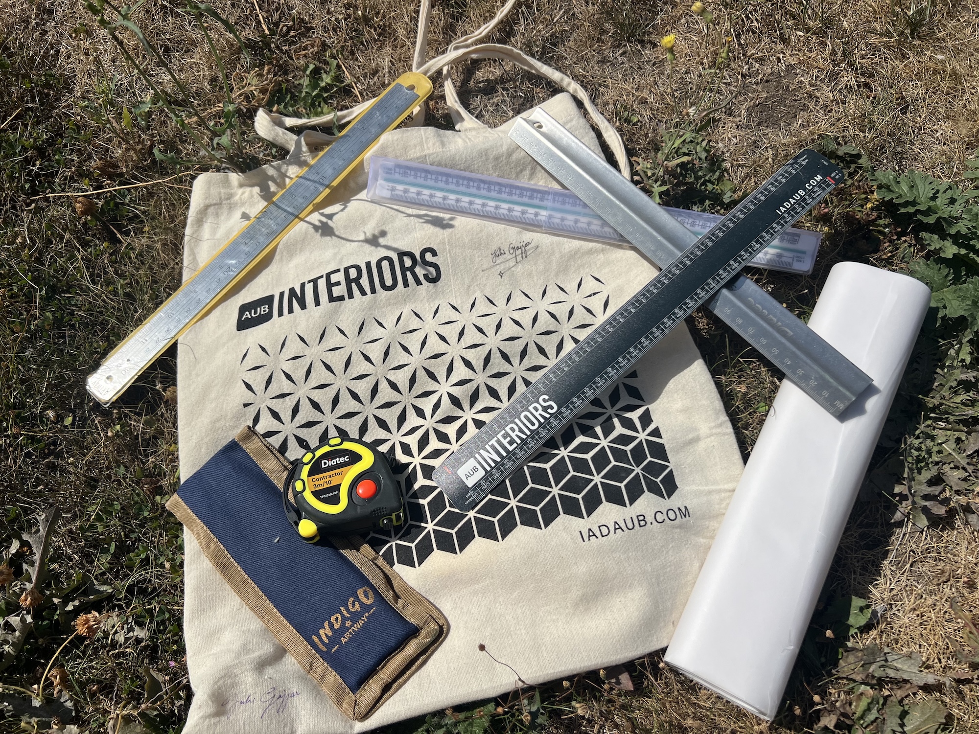 A canvas bag printed with INTERIORS and geometric patterns, surrounded by measuring tools, a tape measure, a pencil case and a roll of white paper on dry grass.