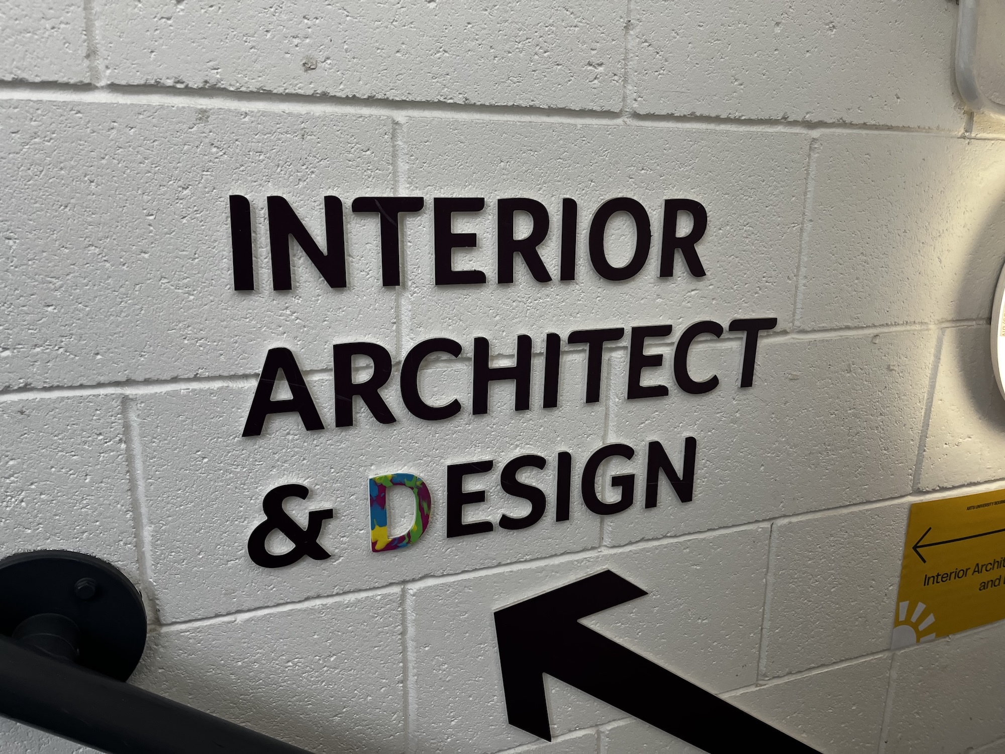 Black text reading "INTERIOR ARCHITECT AND DESIGN" mounted on white painted brick wall with directional arrow pointing left.