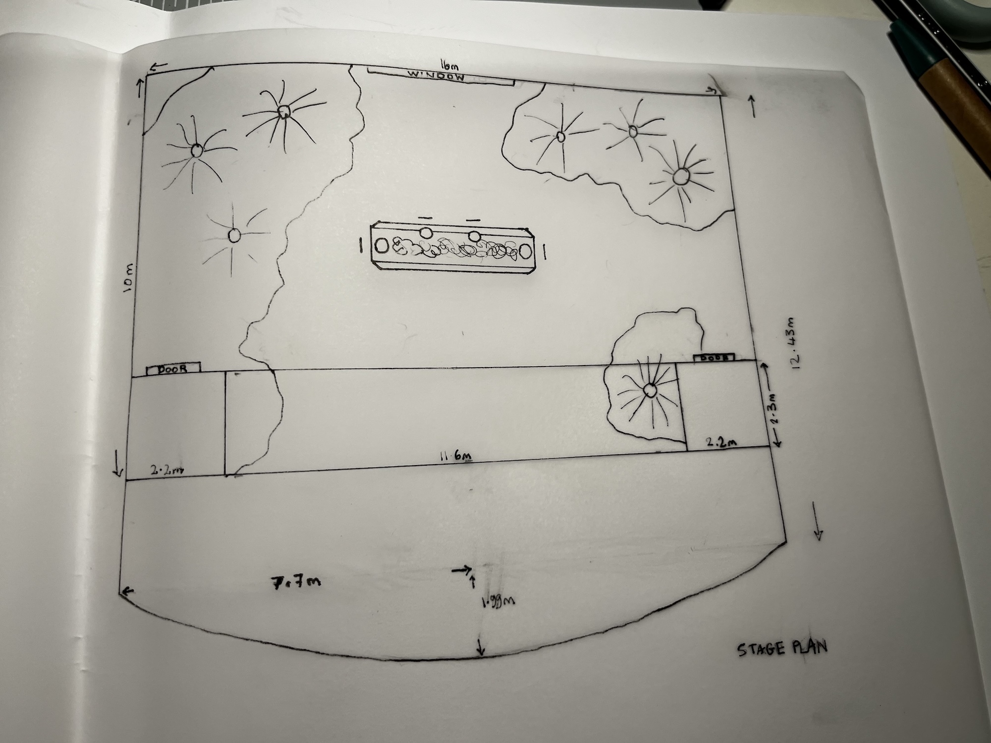 A hand-drawn stage plan on white paper showing a rectangular layout with marked dimensions, flower-like symbols and a central table-like shape, photographed on a desk.