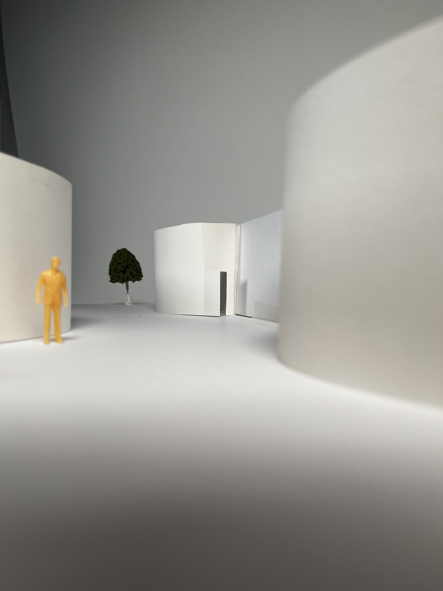 Minimalist architectural rendering with white curved walls, grey surfaces, small yellow figure, and single green tree in bright interior space.