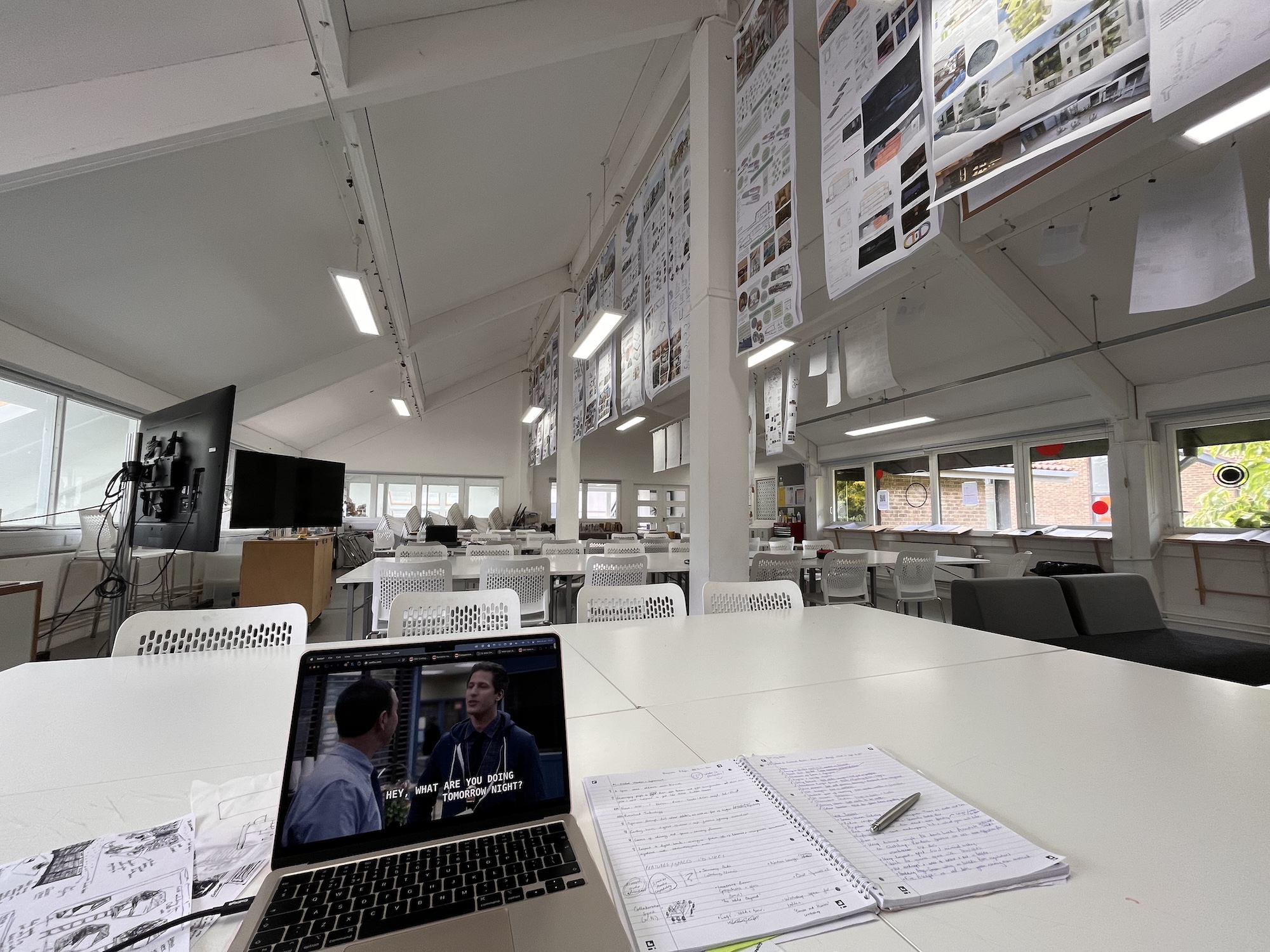 A laptop on a white table displays a video next to an open notebook and pen, in a bright office with rows of desks and posters on the walls.
