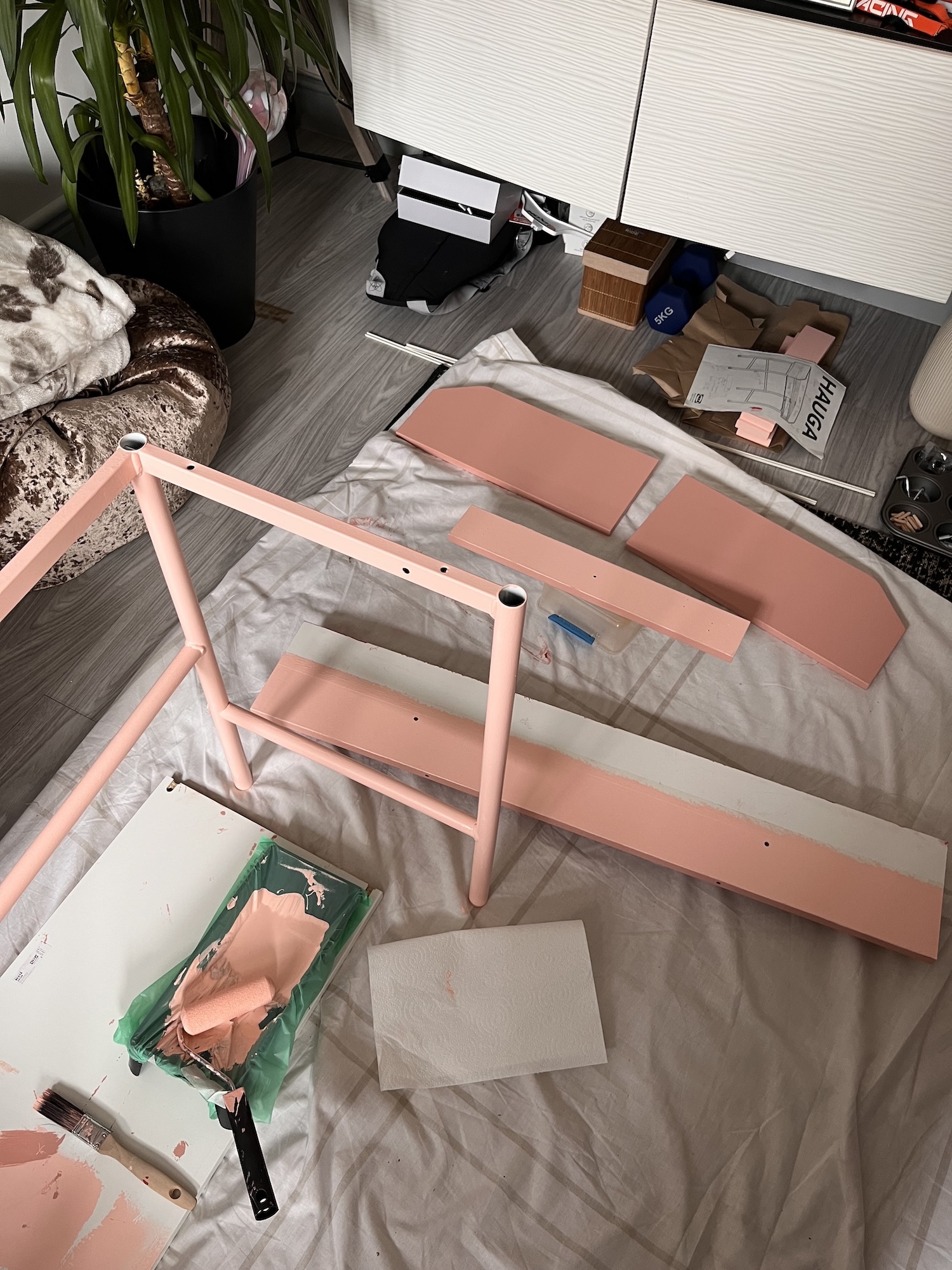 Pink wooden desk frame being painted on protective sheet, with green paint tray and brush nearby on grey floor.