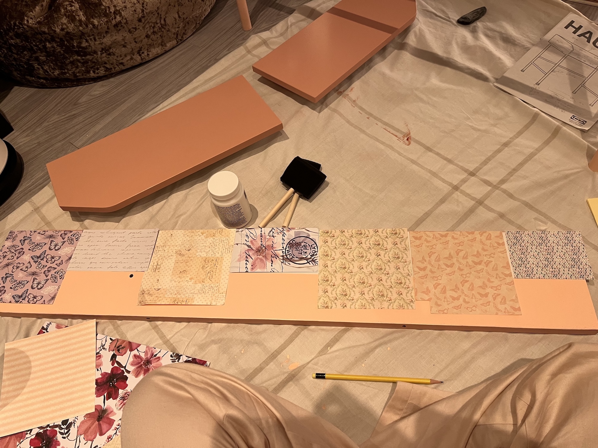 Craft supplies on wooden floor: orange card rectangles, decorative papers with floral patterns, small paint pots, pencil, and woven basket.