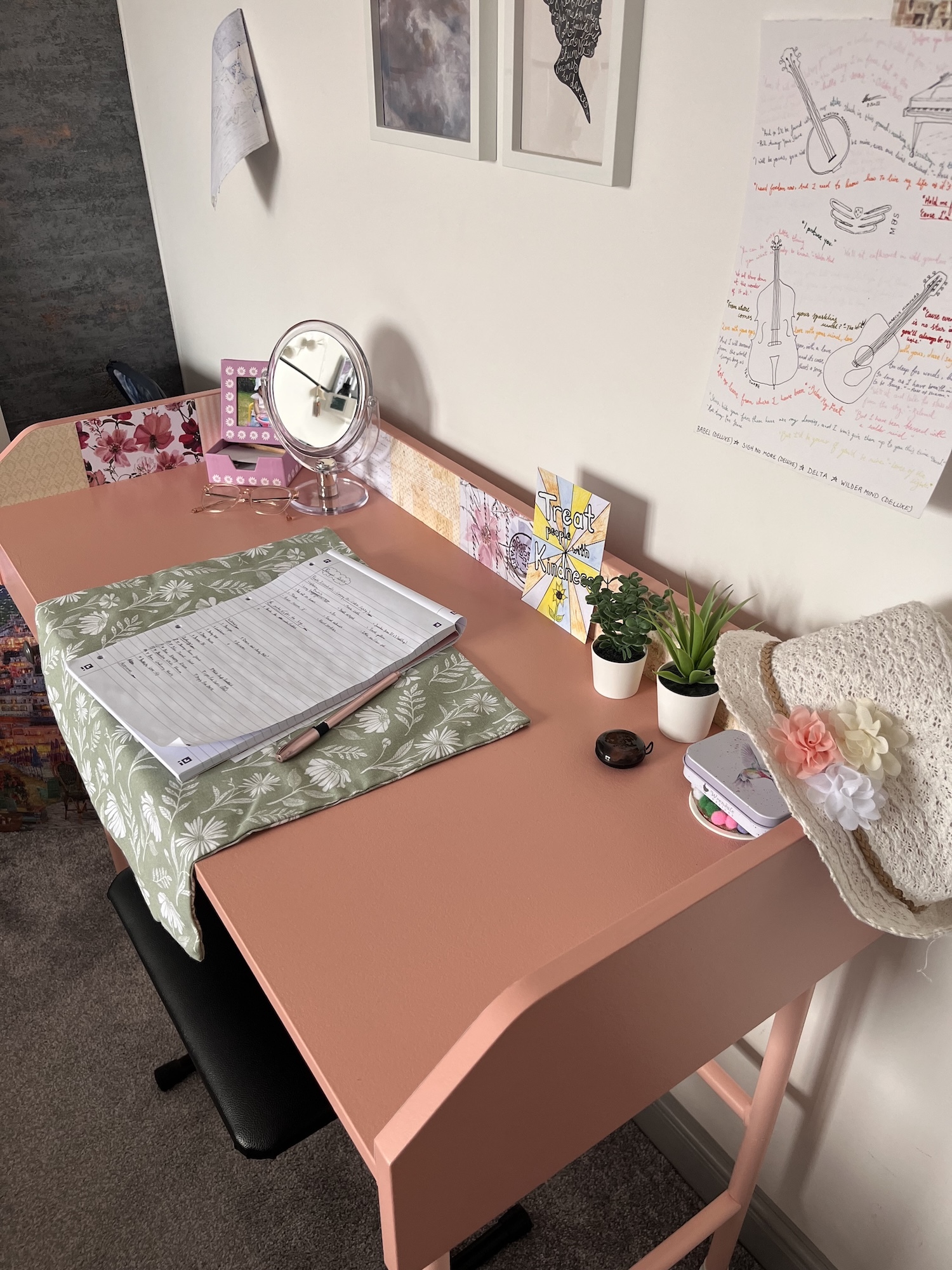 Pink desk with open notebook, round mirror, small plants in pots, and papers on white wall above.