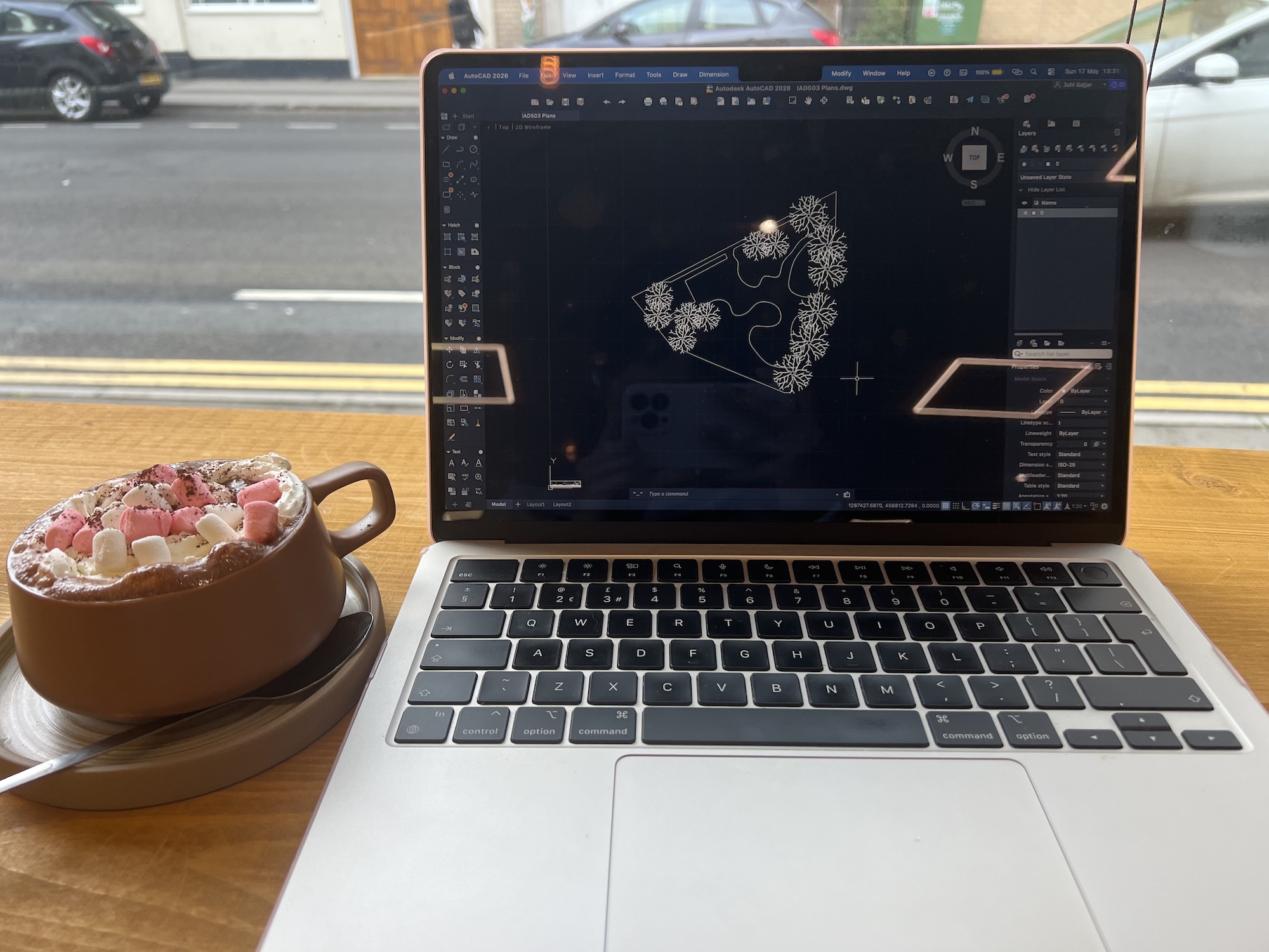 A laptop on a wooden table displays a design interface with a triangular outline and floral shapes, beside a mug topped with cream and pink sprinkles by the window.