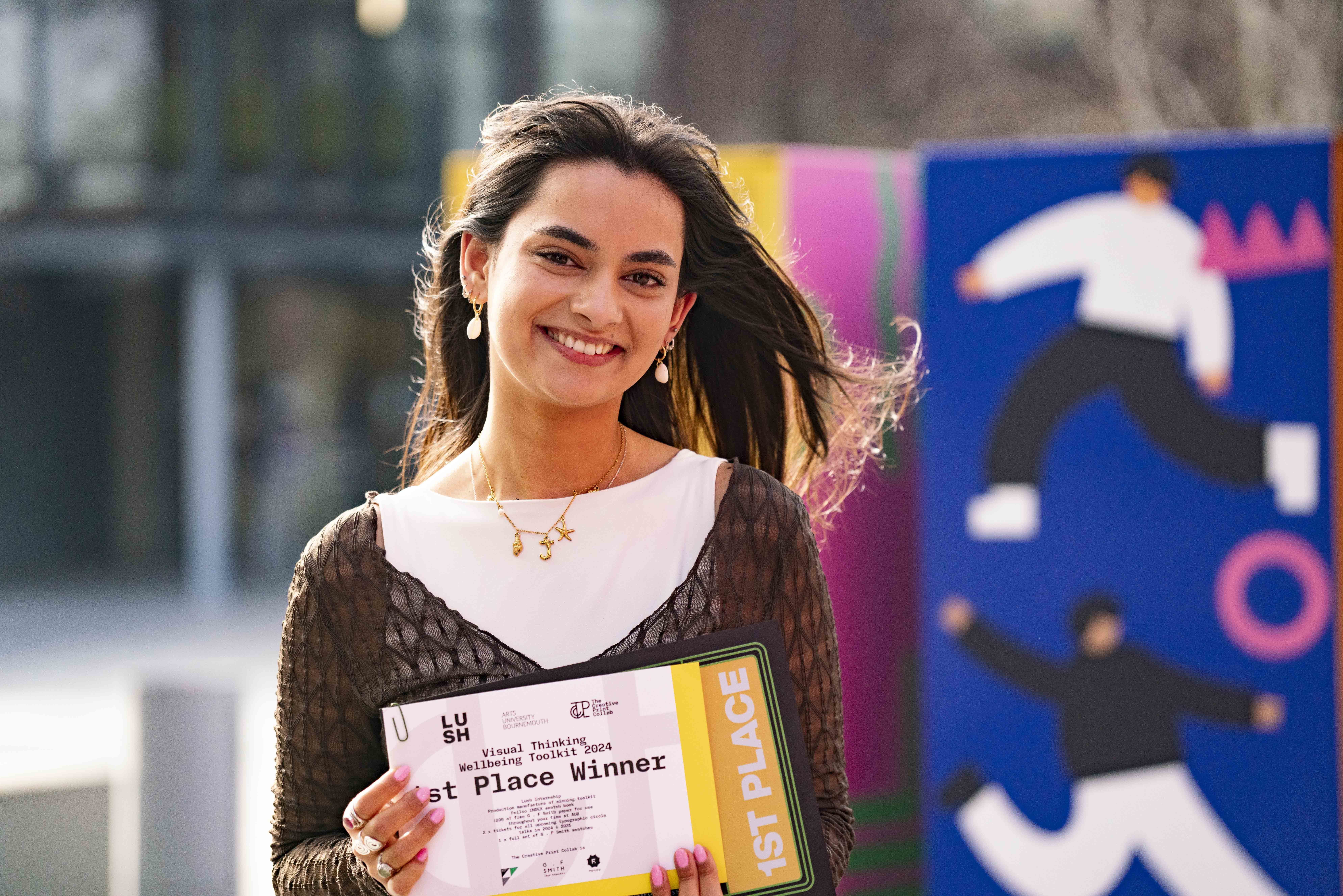 Julia Anderson stood outside on-campus holding a 1st place certificate, blurred background.
