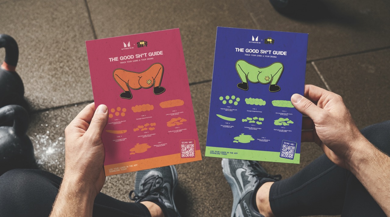 A person holding two colourful brochures titled "The Good Sh*t Guide". One is pink with illustrations and descriptions, while the other is blue, also featuring illustrations and text. A kettlebell is visible in the background on a gym floor.