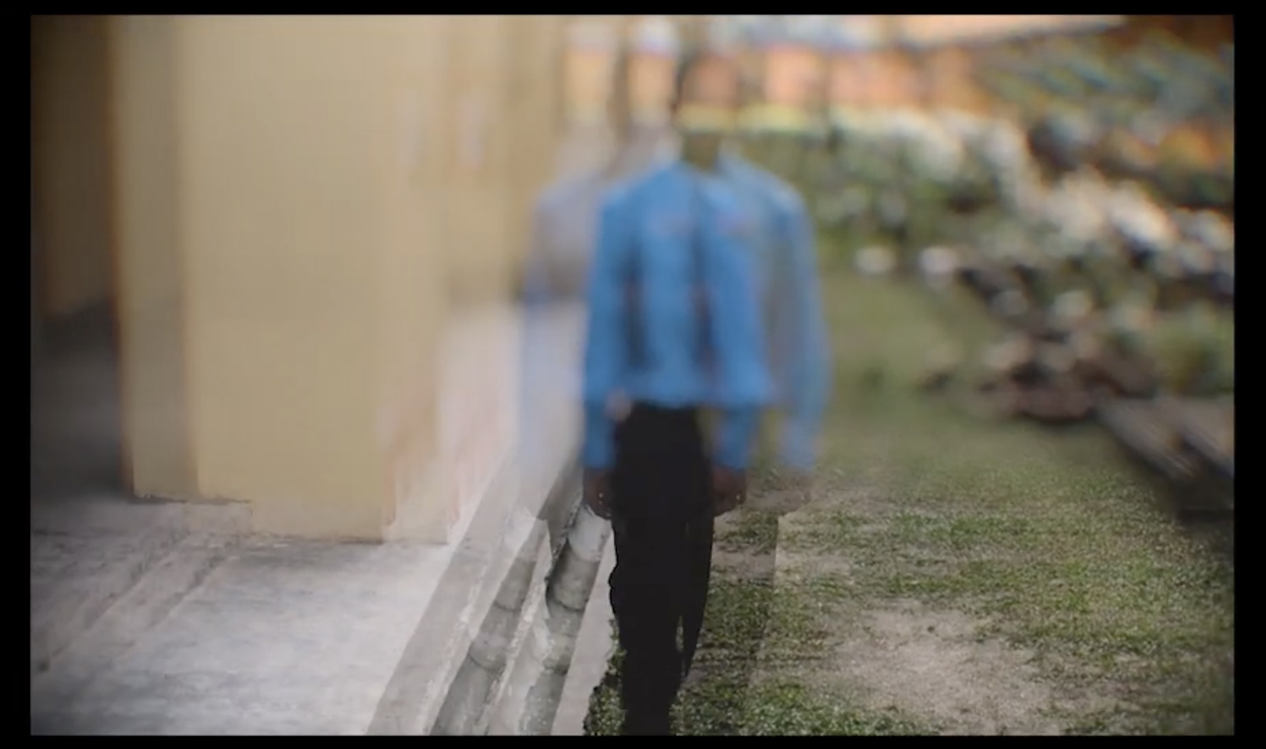Blurred figure in blue shirt walking away down outdoor path with stone wall and greenery visible to the right.