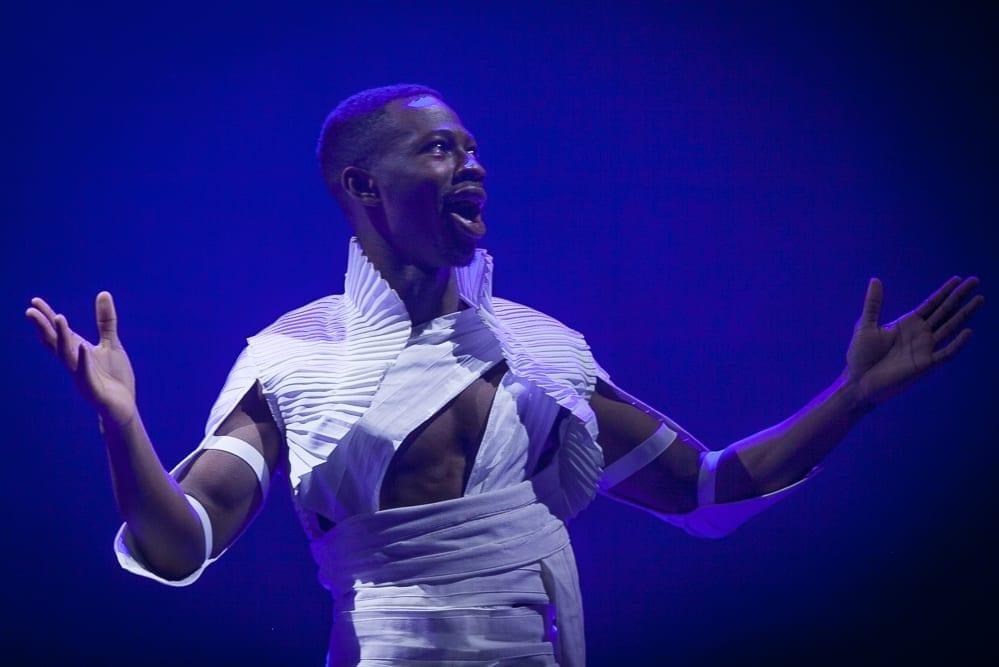 Actor in costume with his arms raised, on stage in mid-speech.
