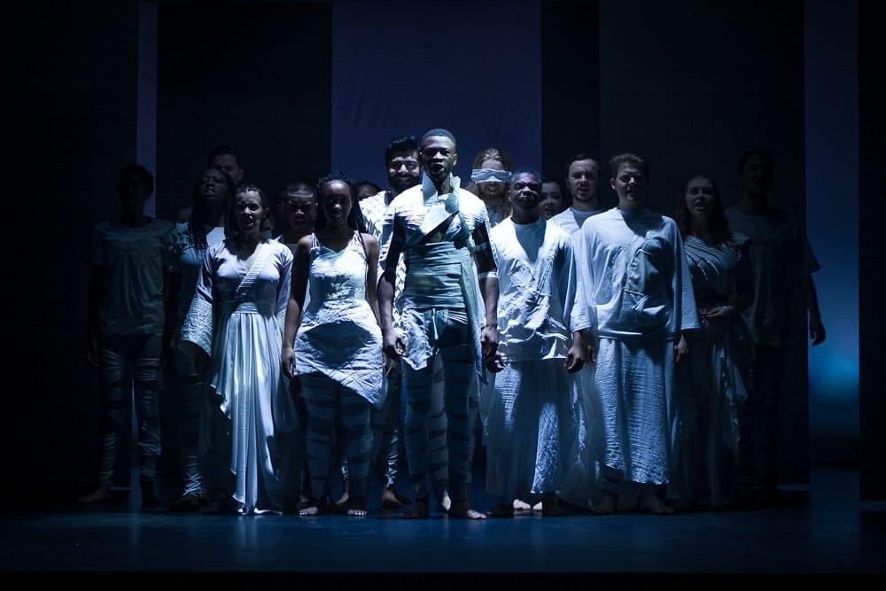 A large group of performers in costume underneath a spotlight on the stage.