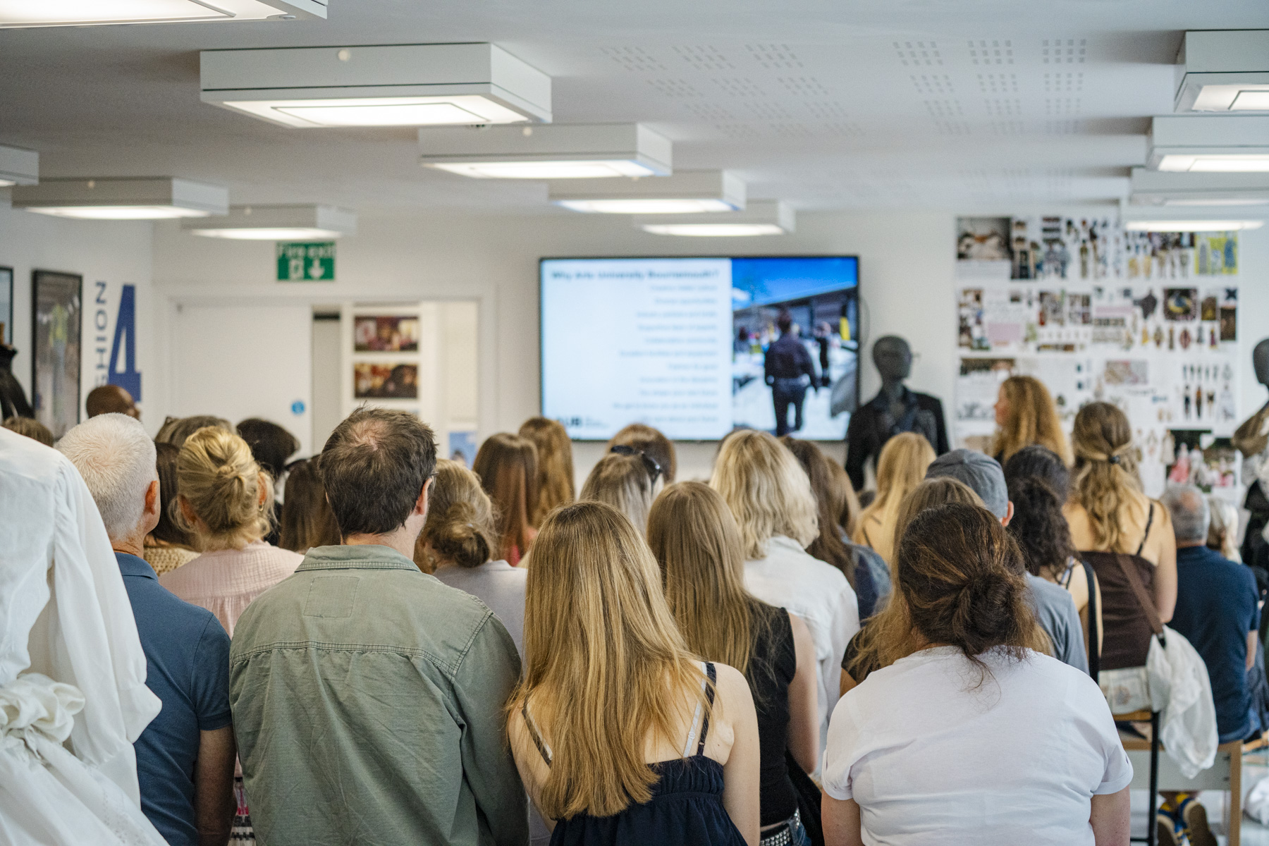 An audience of seated and standing people faces a presentation screen in a bright room with fluorescent ceiling lights and wall displays.